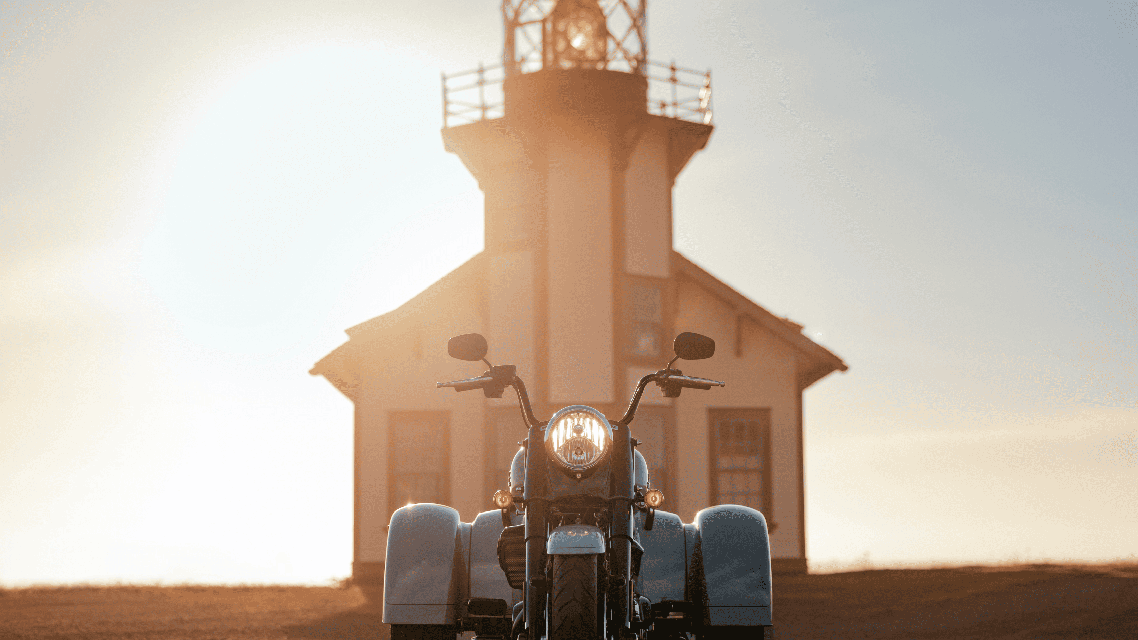 Front view of the 2024 Harley-Davidson® Freewheeler® illuminated by soft sunlight, parked in front of a rustic lighthouse, capturing its bold design and adventurous spirit