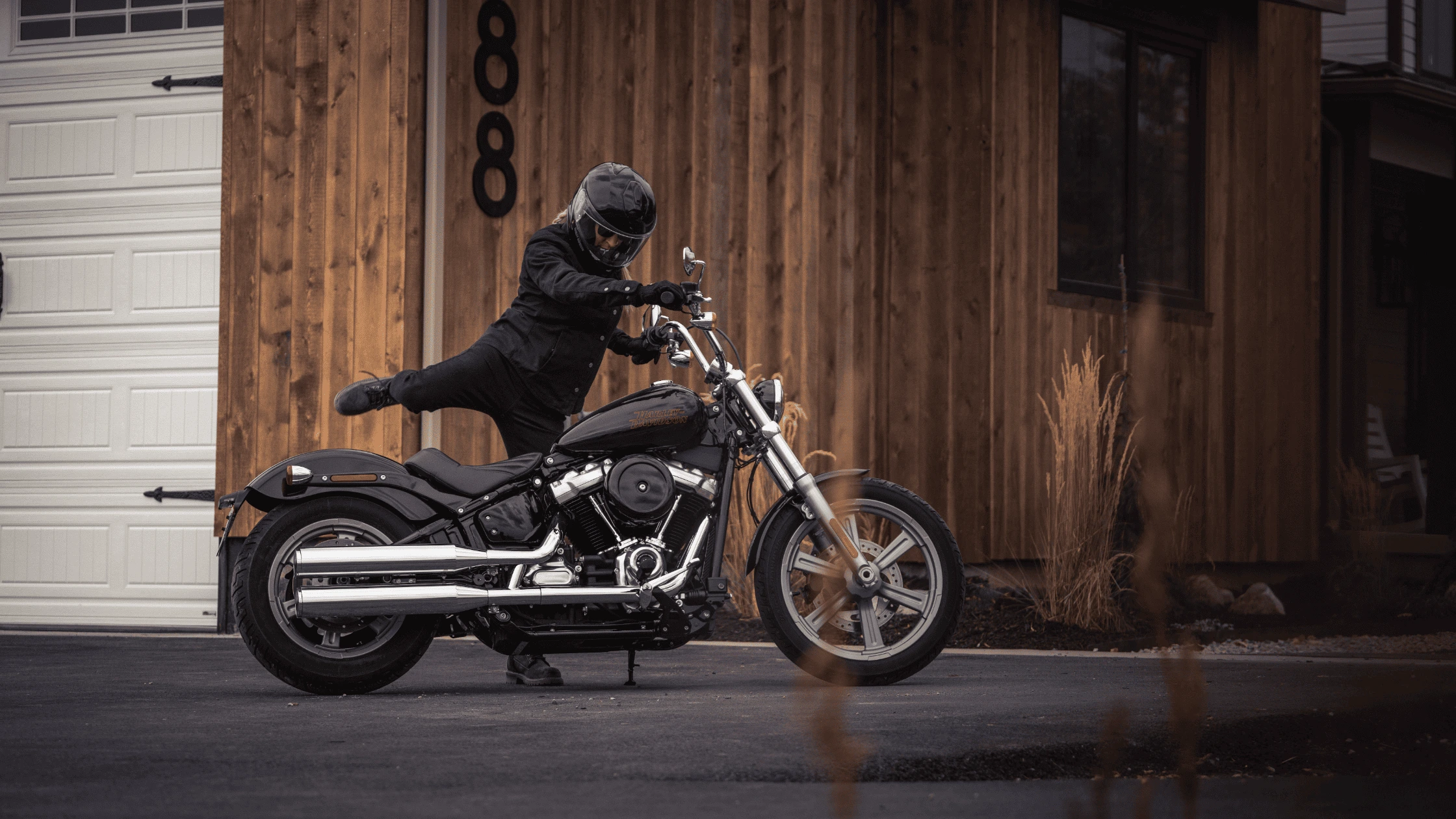 A rider mounting the 2024 Harley-Davidson® Softail® Standard near Bentonville, AR, in front of a rustic wooden building, emphasizing the bike's classic cruiser style.