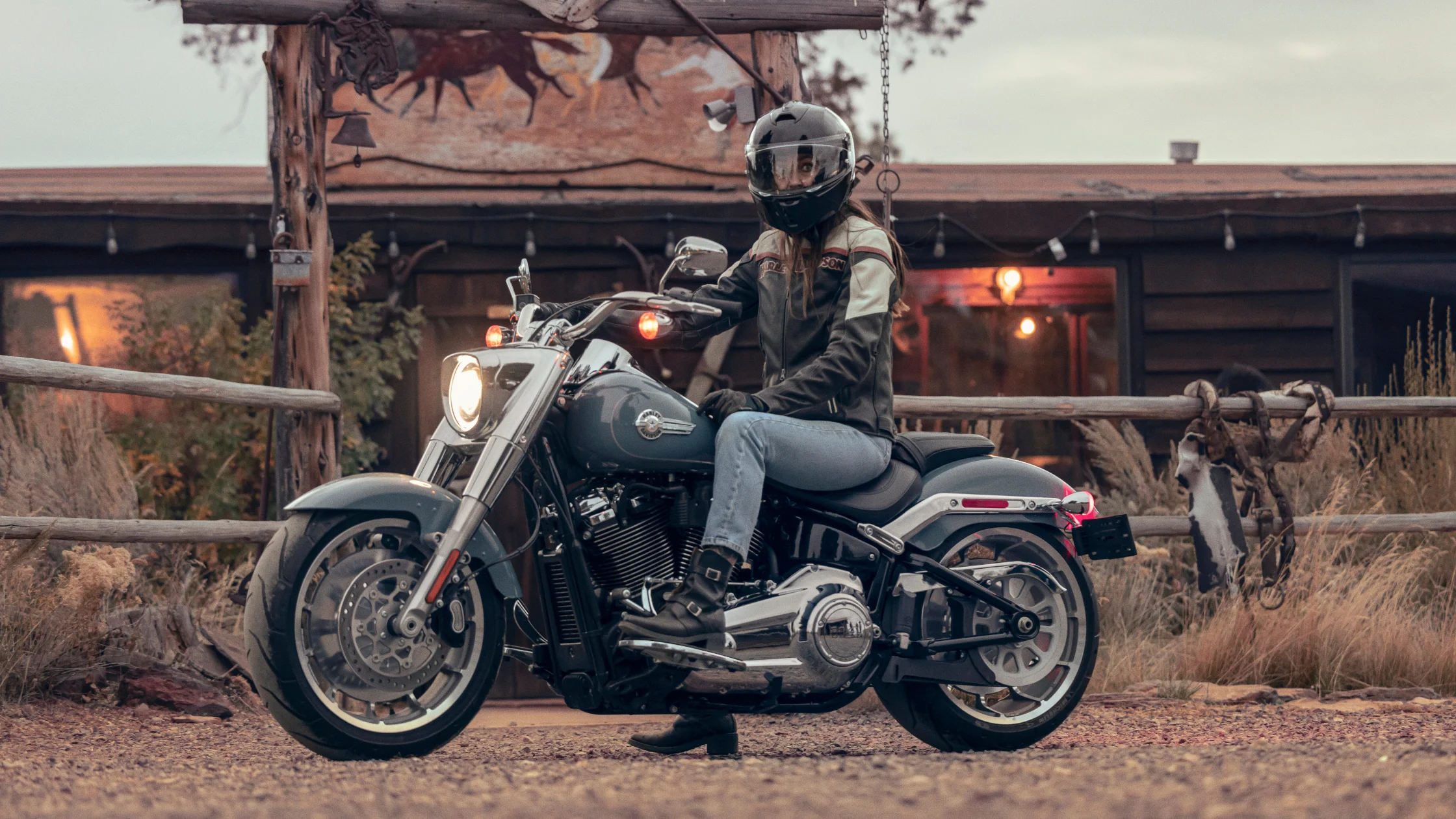 A rider on the 2024 Harley-Davidson® Fat Boy® 114 parked near a rustic cabin.