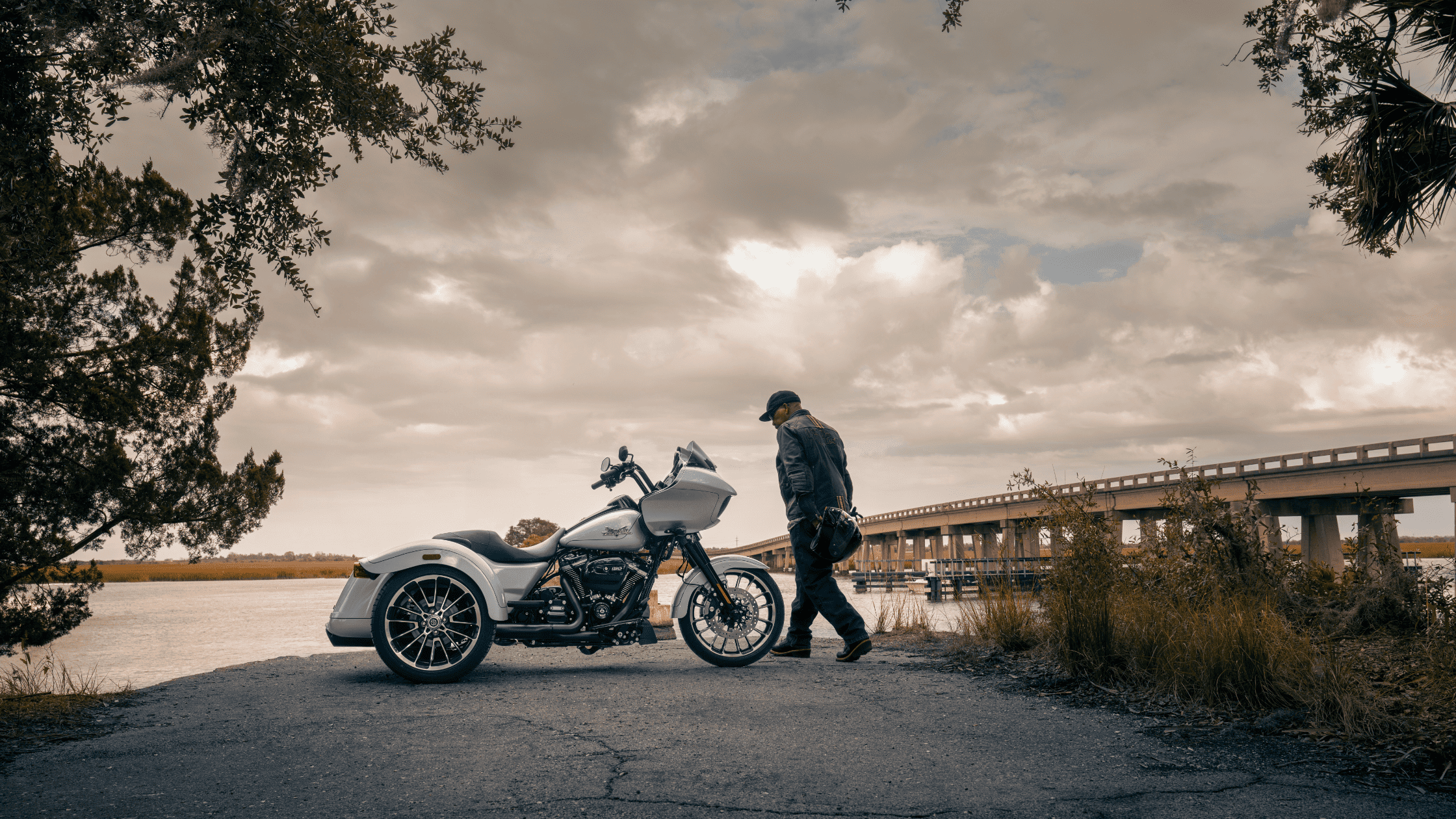 A man stands next to a Harley Davidson motorcycle on a road, showcasing his love for biking and the outdoors.