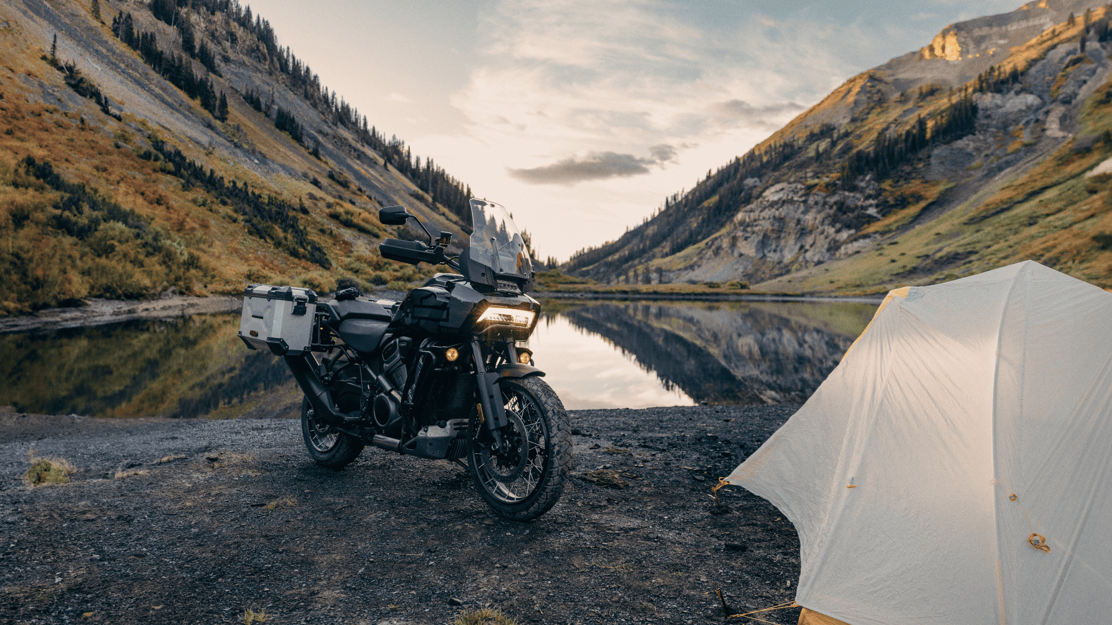 A Harley Davidson motorcycle parked beside a tent near a serene lake, surrounded by nature.