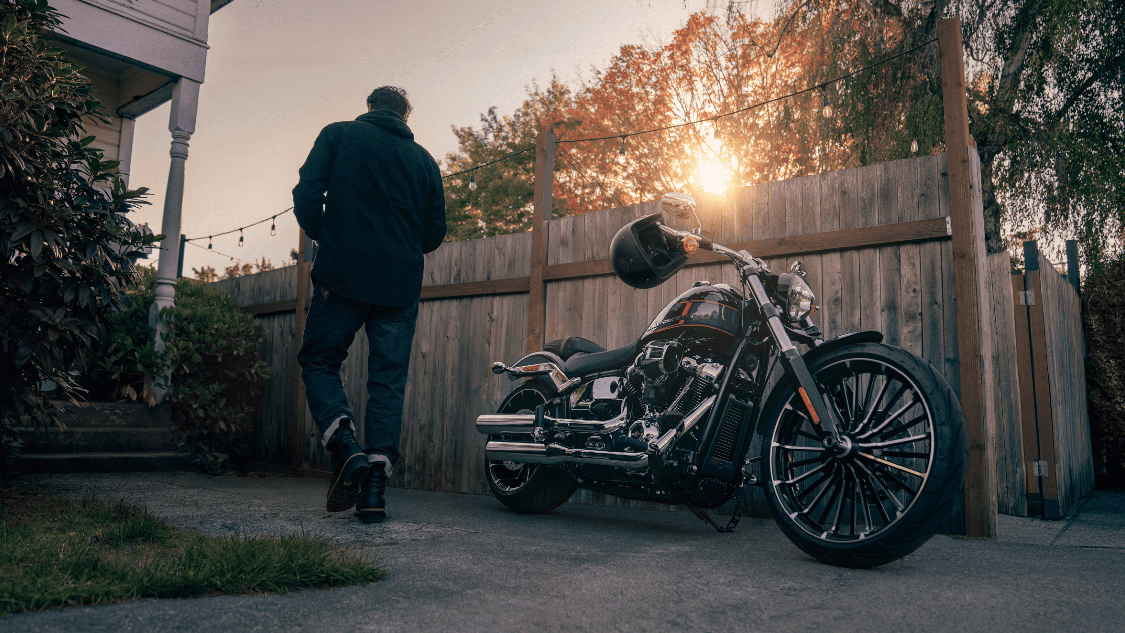 A man walks past a parked Harley Davidson motorcycle in a driveway.