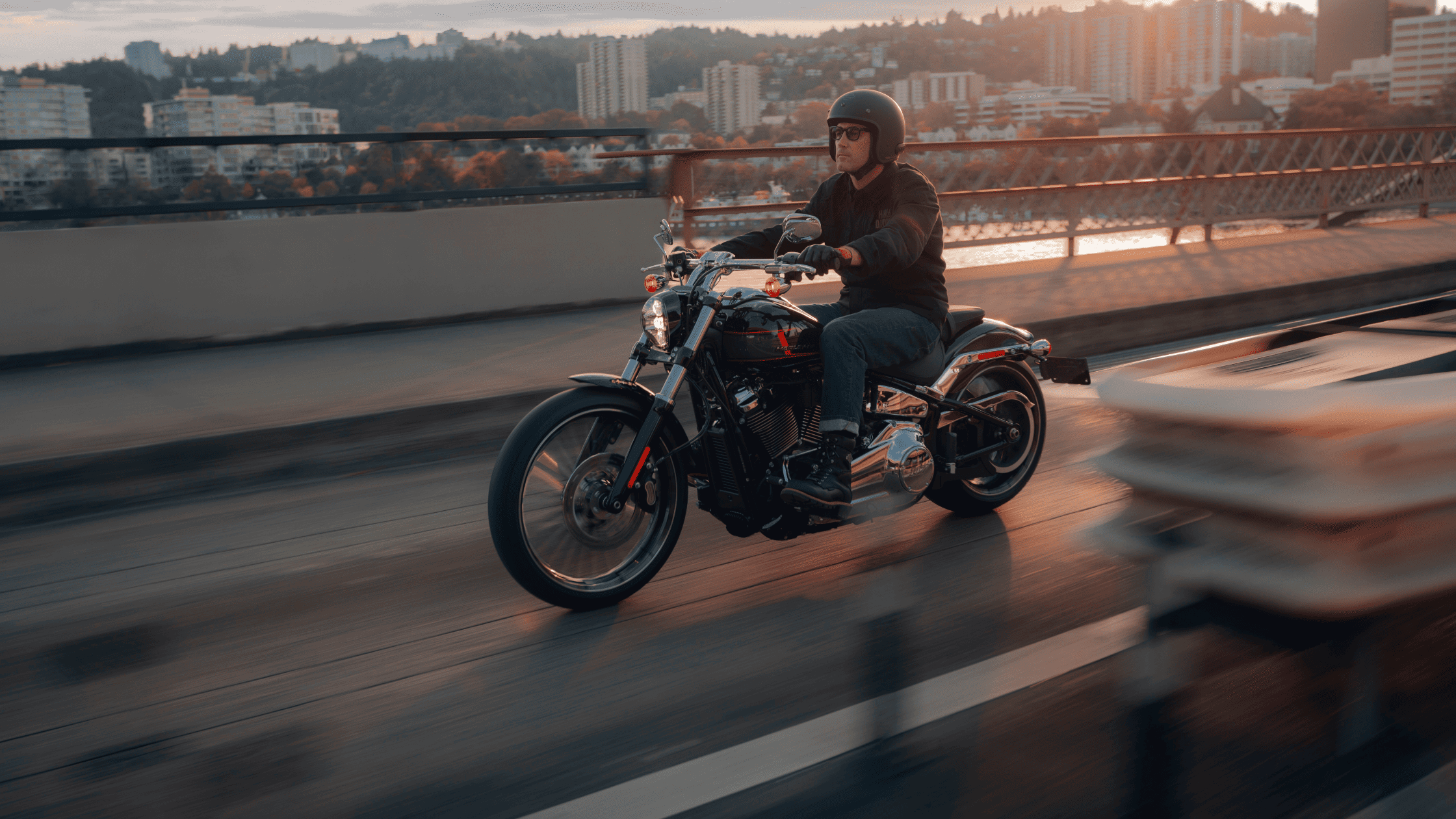 A rider cruising on a 2024 Harley-Davidson® Breakout® 117 across a scenic bridge during sunset