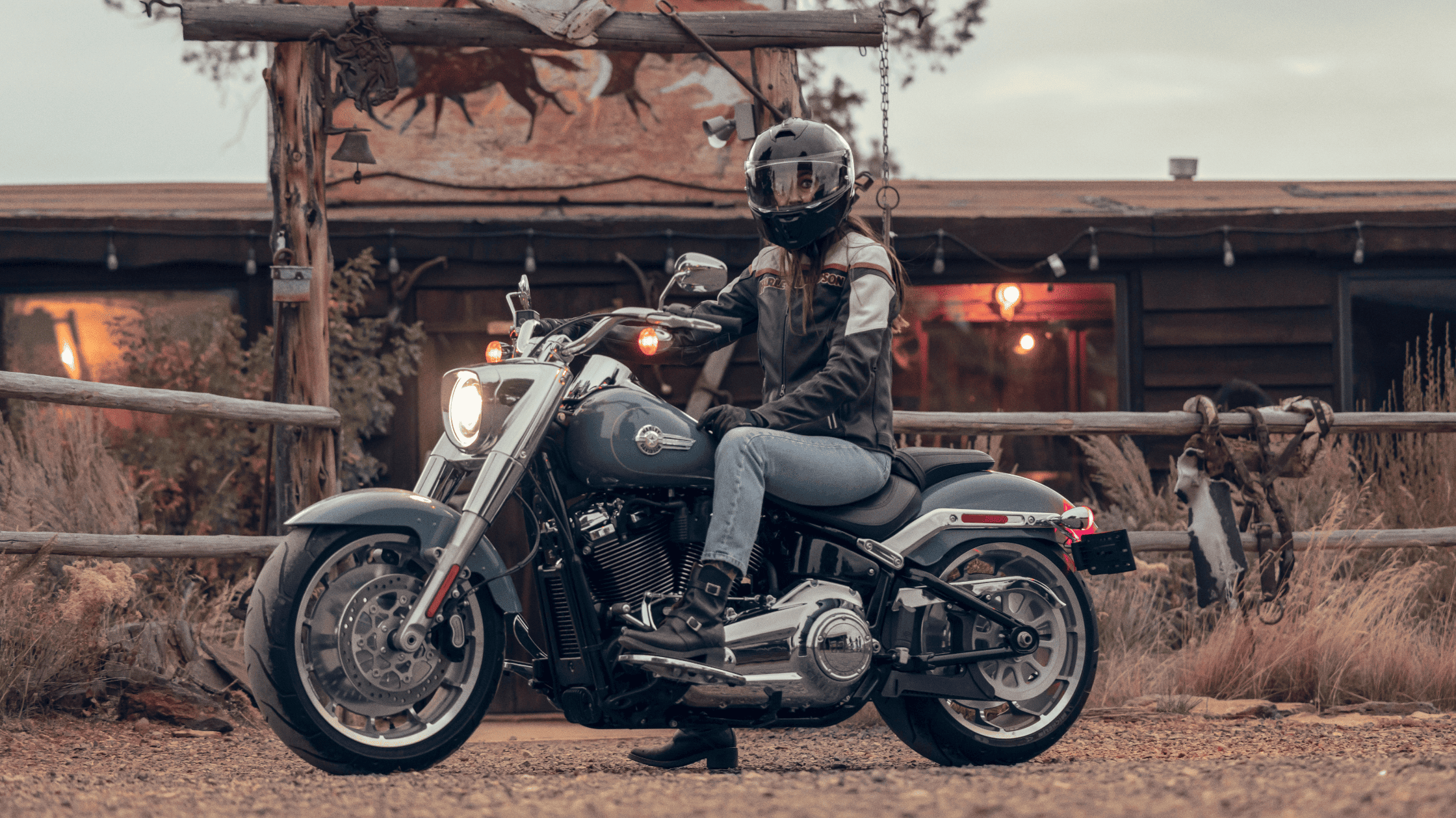 A rider on a 2024 Harley-Davidson® Fat Boy® 114 parked in front of a rustic cabin with warm lighting.