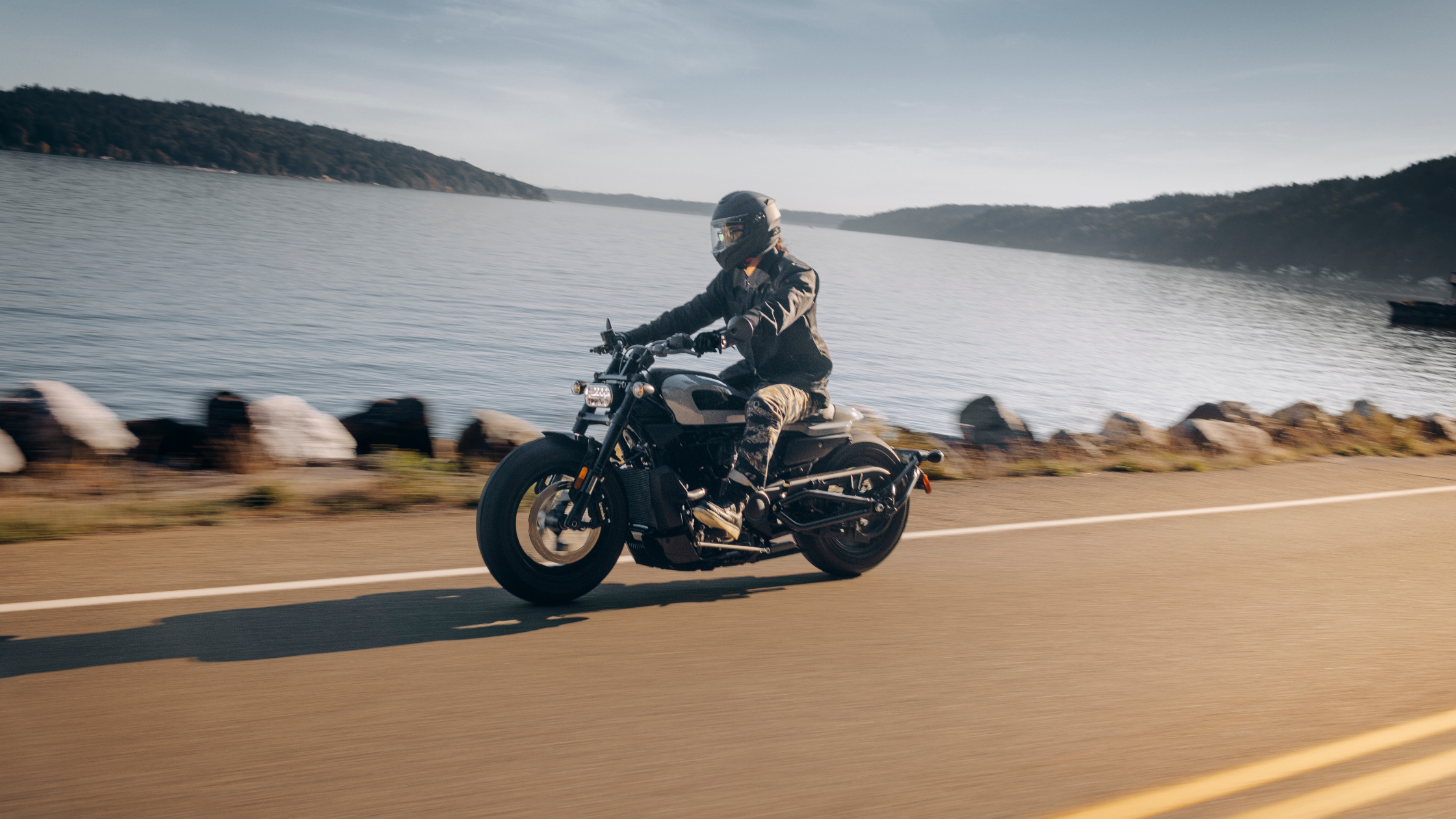 A rider on a 2024 Harley-Davidson® Sportster® S cruises along a scenic road beside shimmering water.