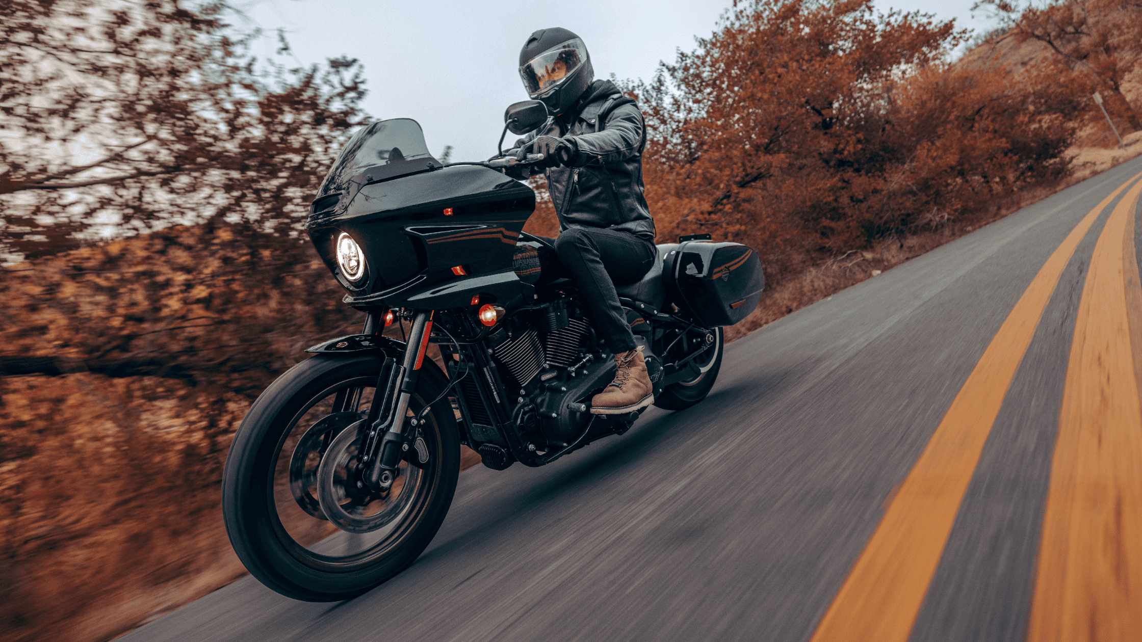 A rider on a 2024 Harley-Davidson® Low Rider® ST cruises on a scenic autumn road, wearing full gear