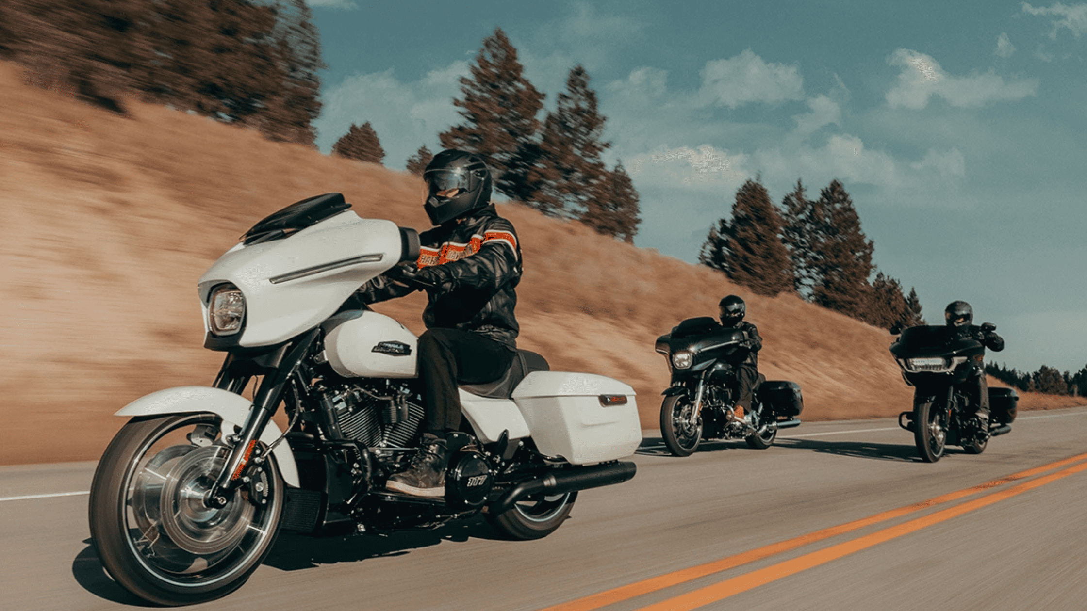Three riders on touring motorcycles, led by a white bike, cruising a scenic road.