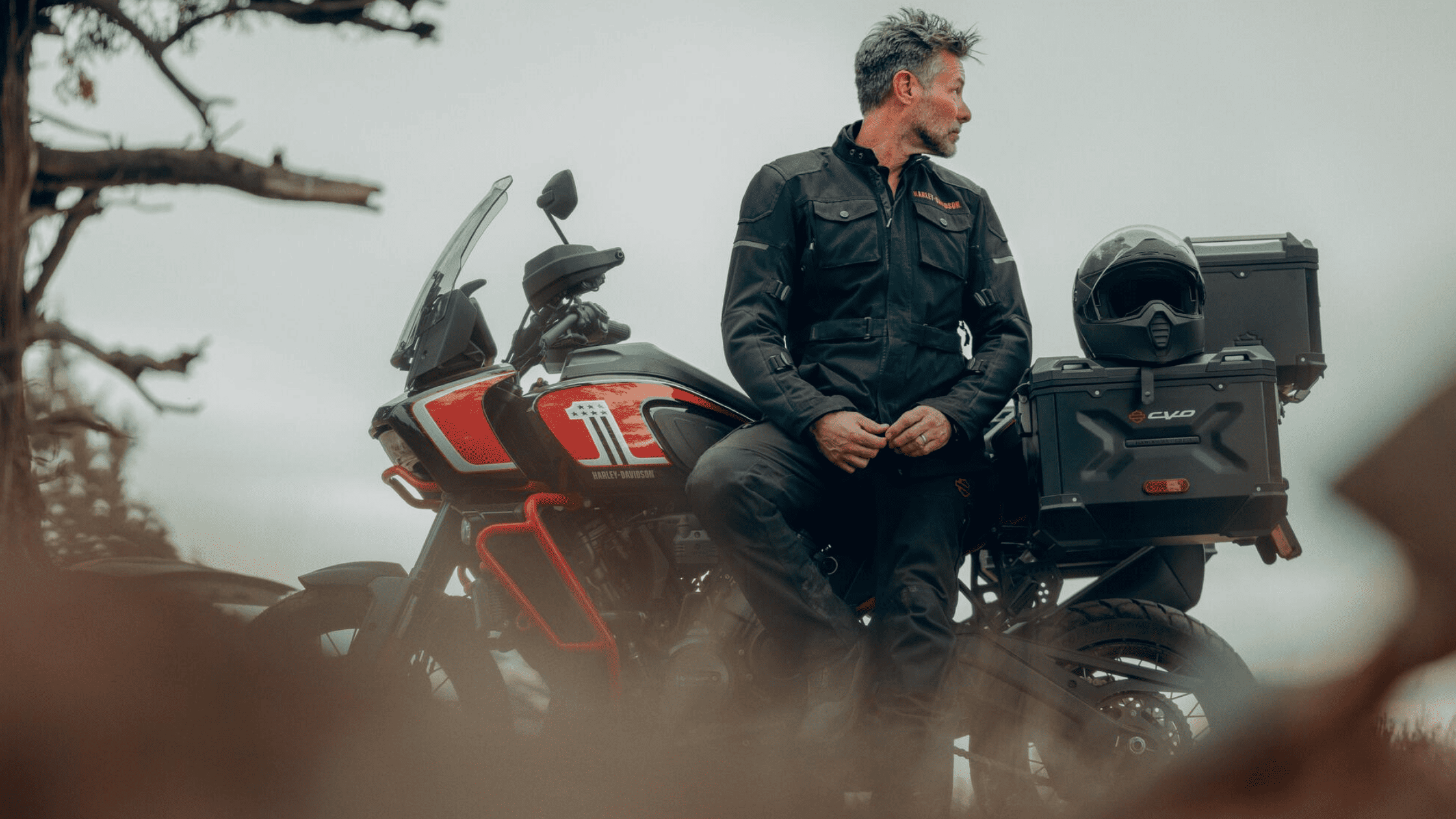 Rider in a black jacket leaning on a red adventure motorcycle with gear storage.