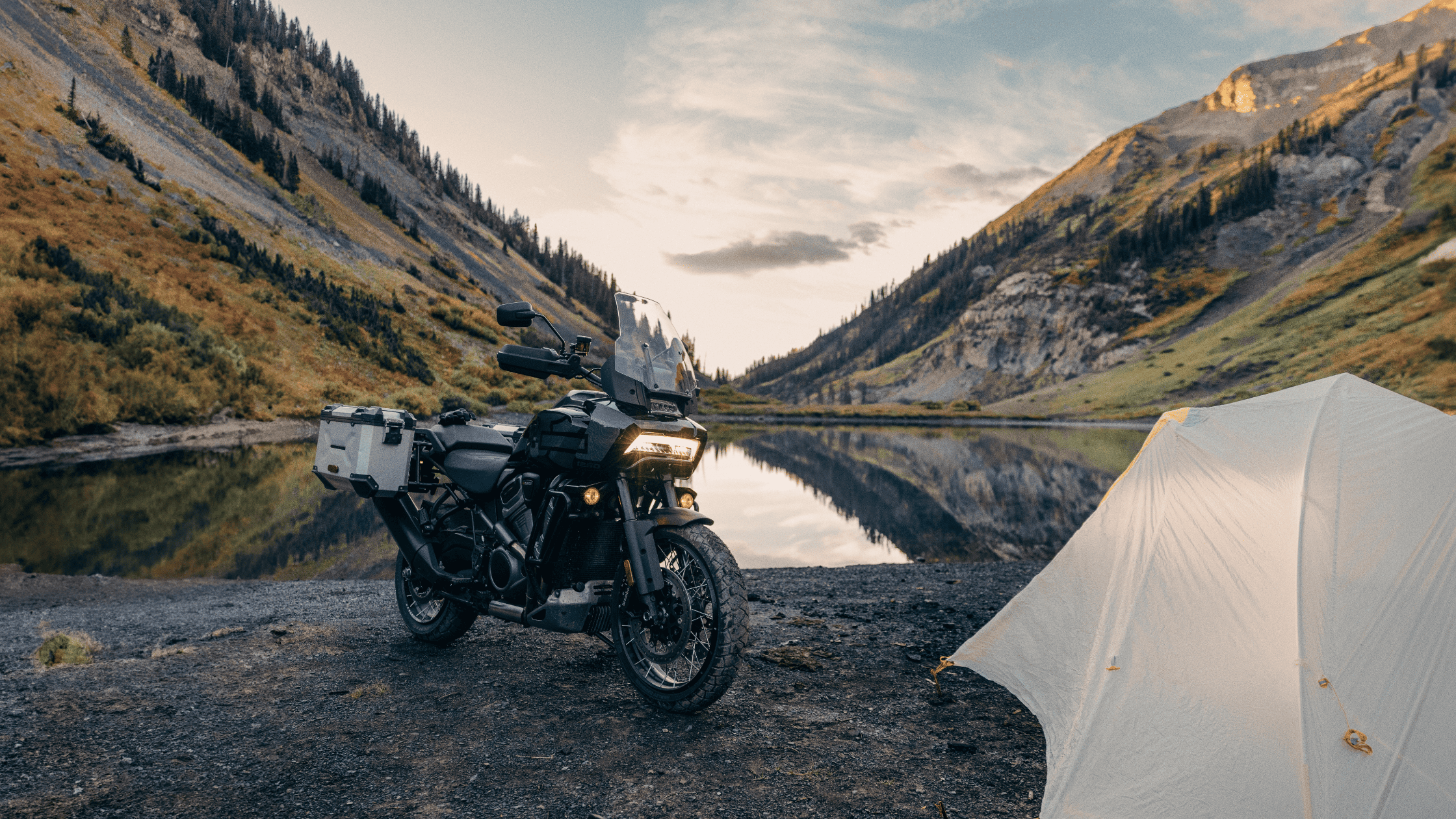 Adventure motorcycle parked by a serene mountain lake with a tent nearby.