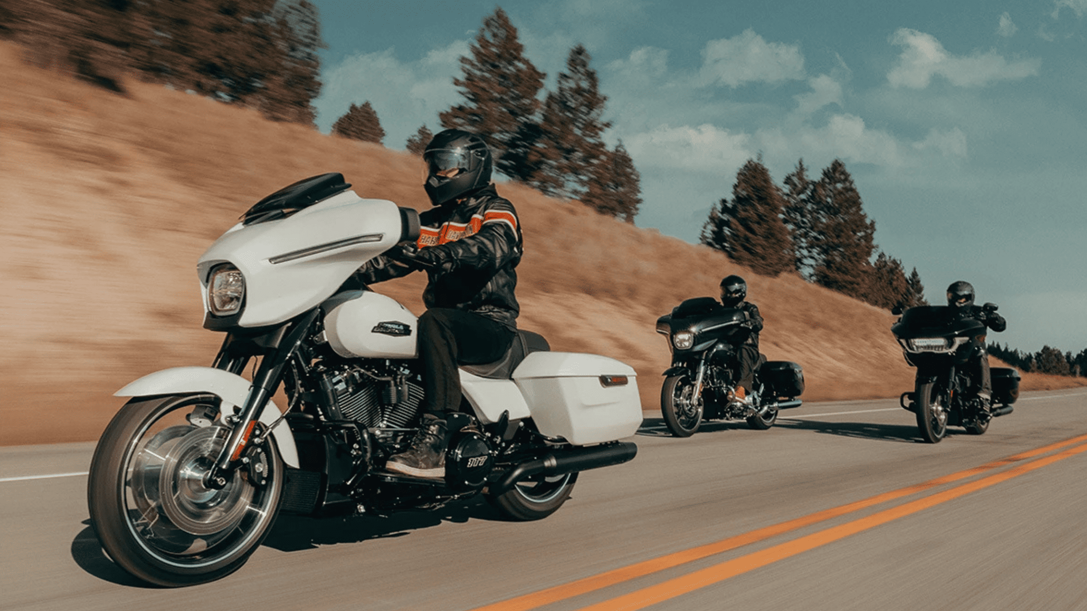Three riders on touring motorcycles, led by a white bike, cruising a scenic road.