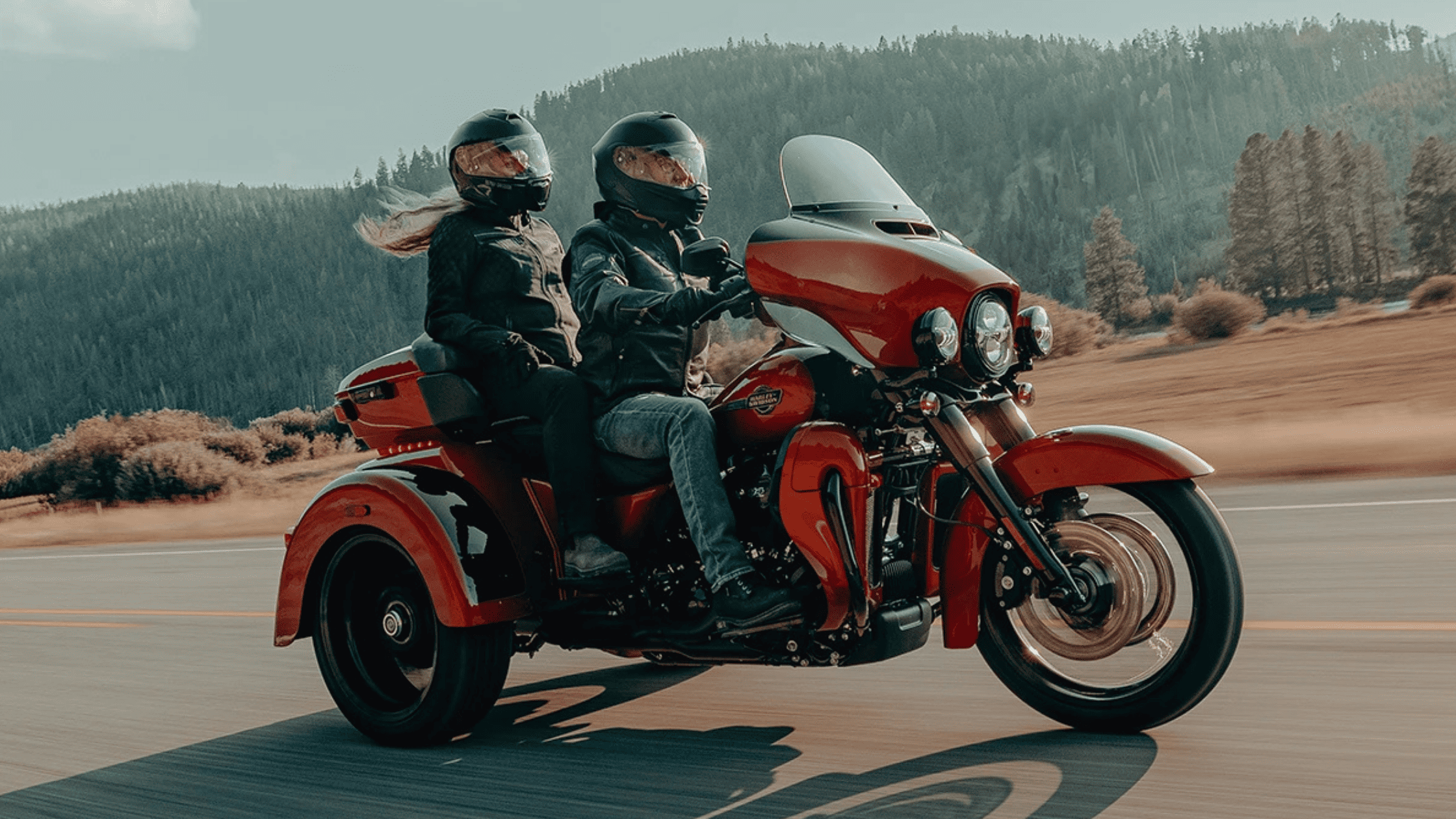 Two riders on a red trike motorcycle cruising a scenic highway.