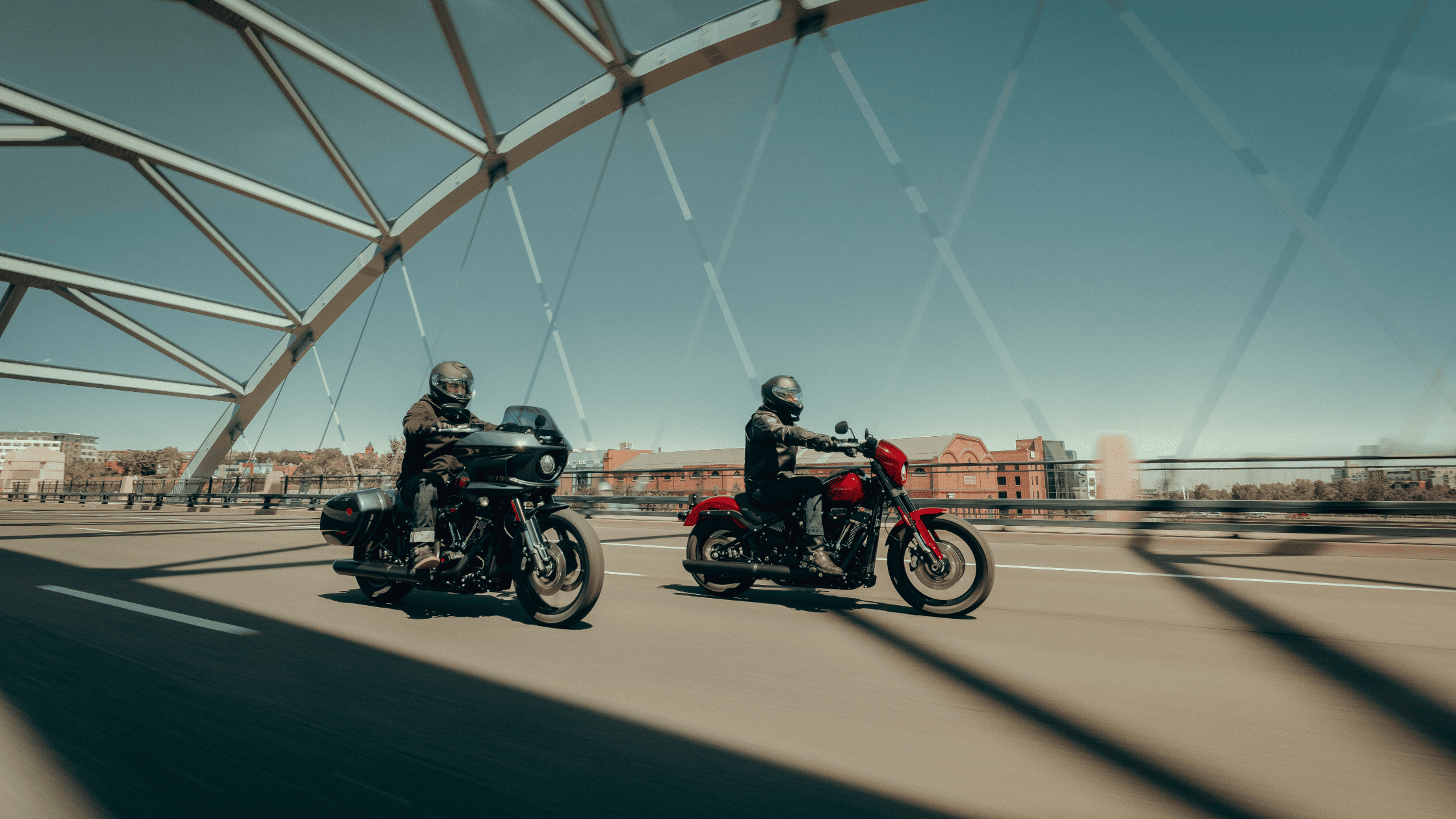 Two motorcyclists riding across a modern bridge in daylight.
