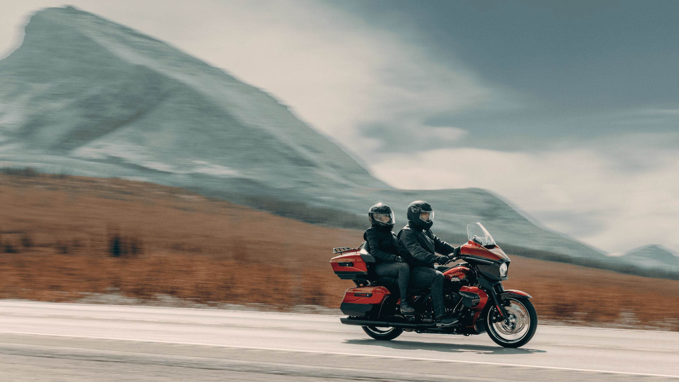 Two riders on a red touring motorcycle with mountain backdrop.