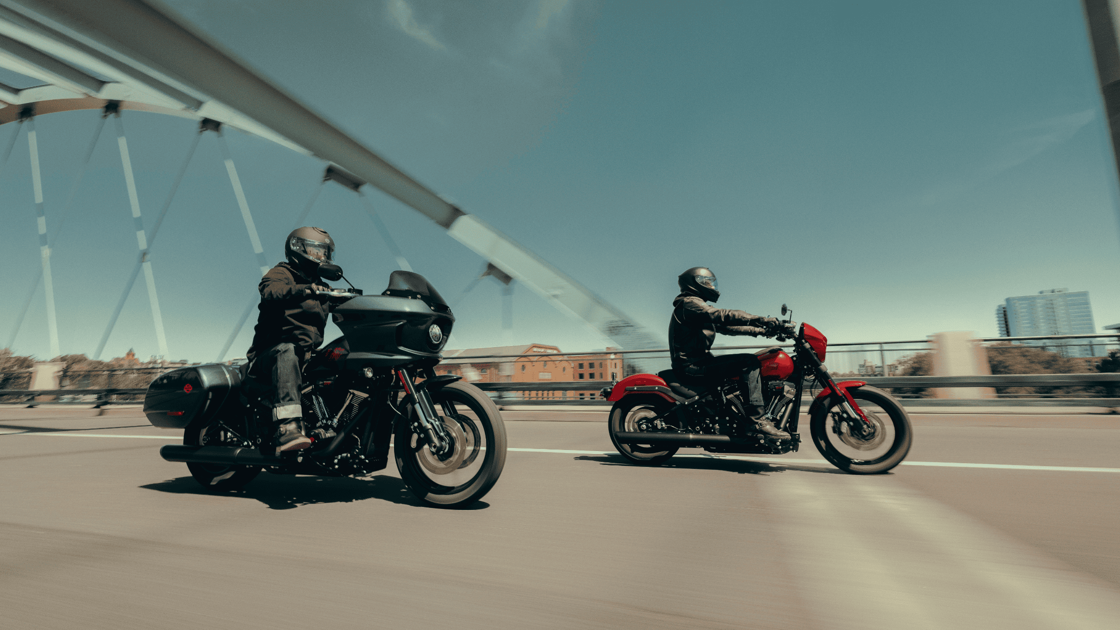 Two motorcyclists riding across a modern bridge in daylight.