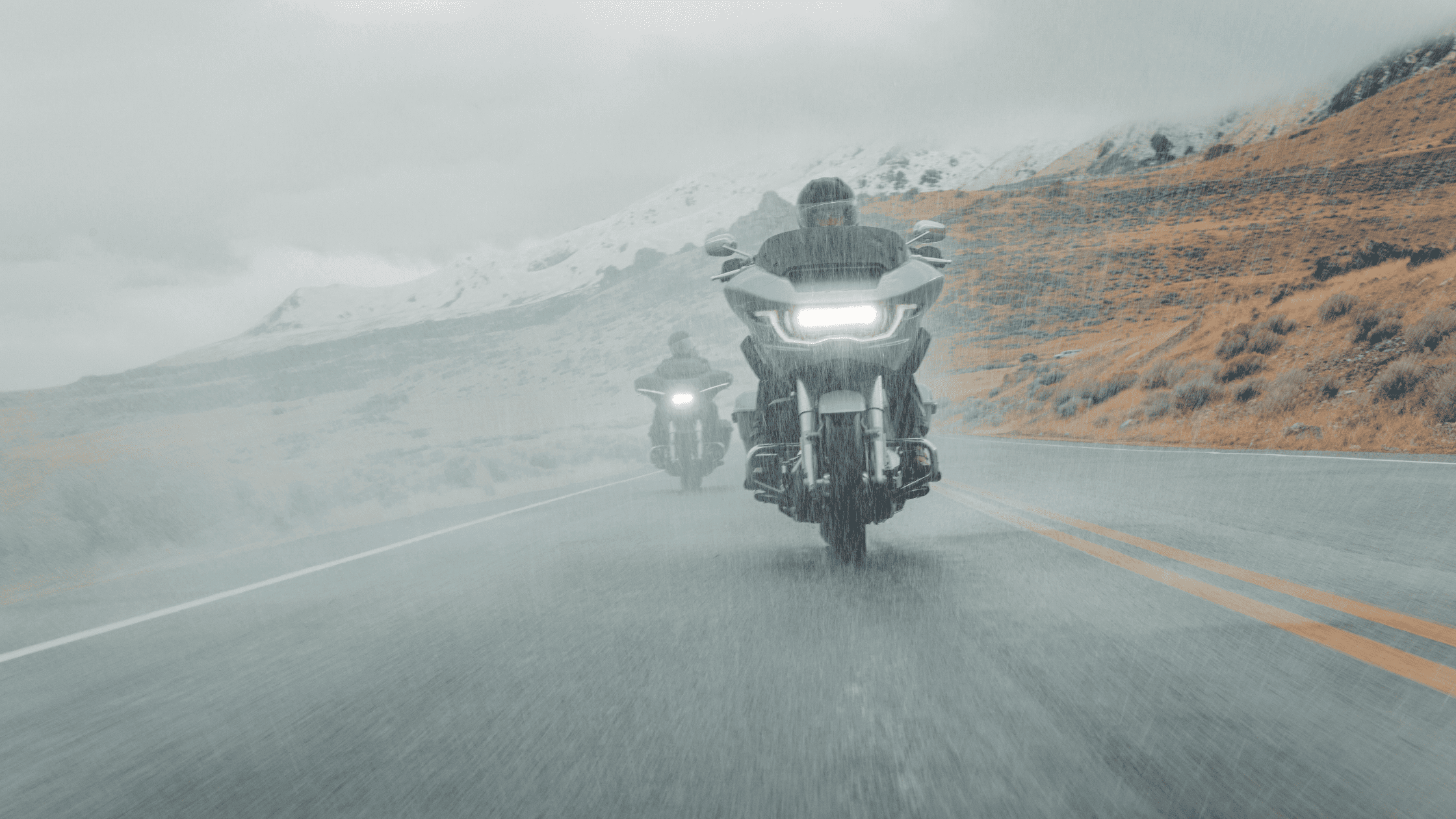 Two motorcyclists ride through a rainy mountain road, with headlights on and misty peaks in the background