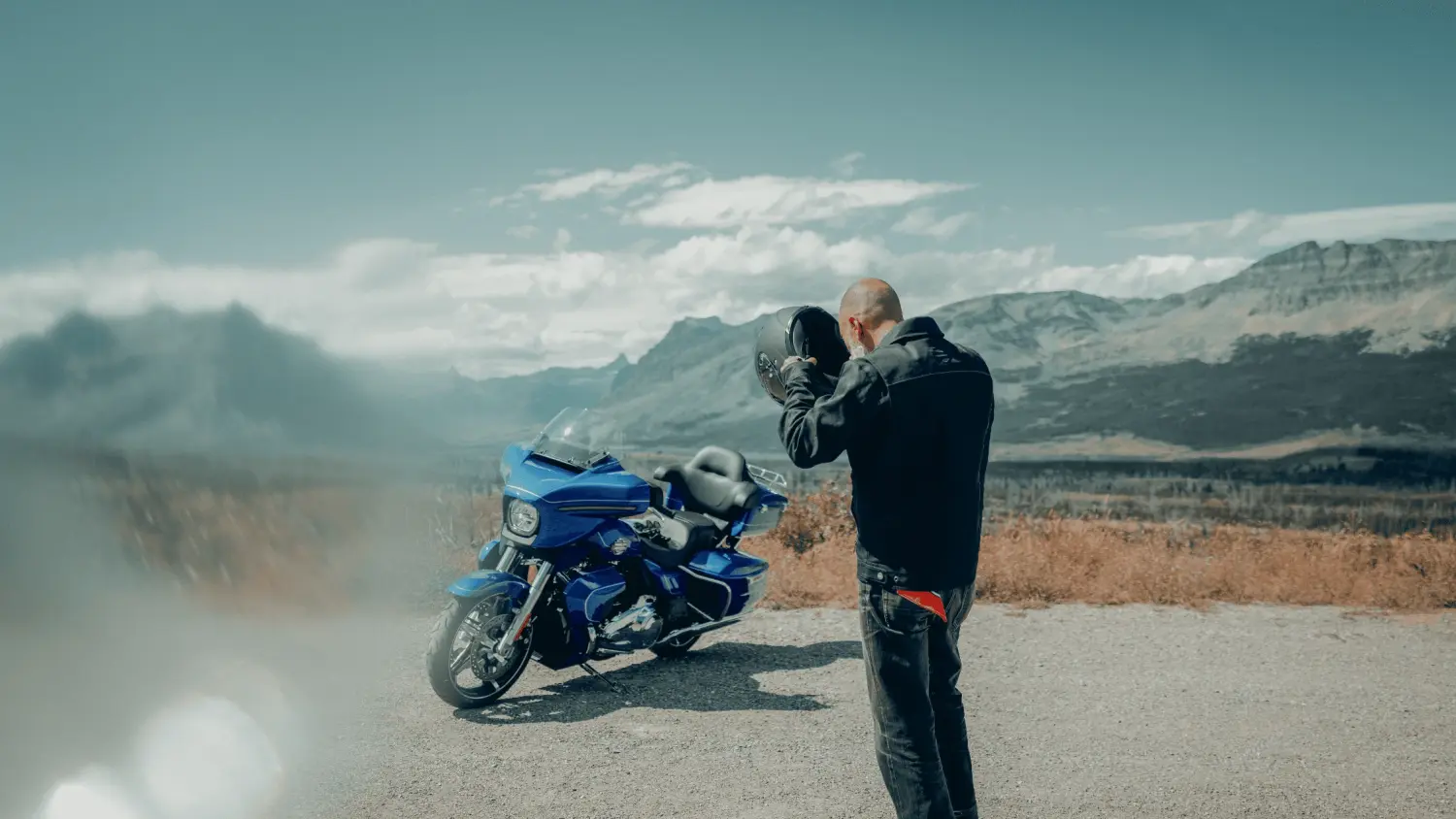 A motorcyclist stands on a scenic overlook, holding a helmet in both hands while gazing at a blue touring motorcycle parked nearby. The backdrop features a breathtaking mountain landscape with rugged peaks and a partly cloudy sky. The image captures the essence of open-road adventure and freedom.