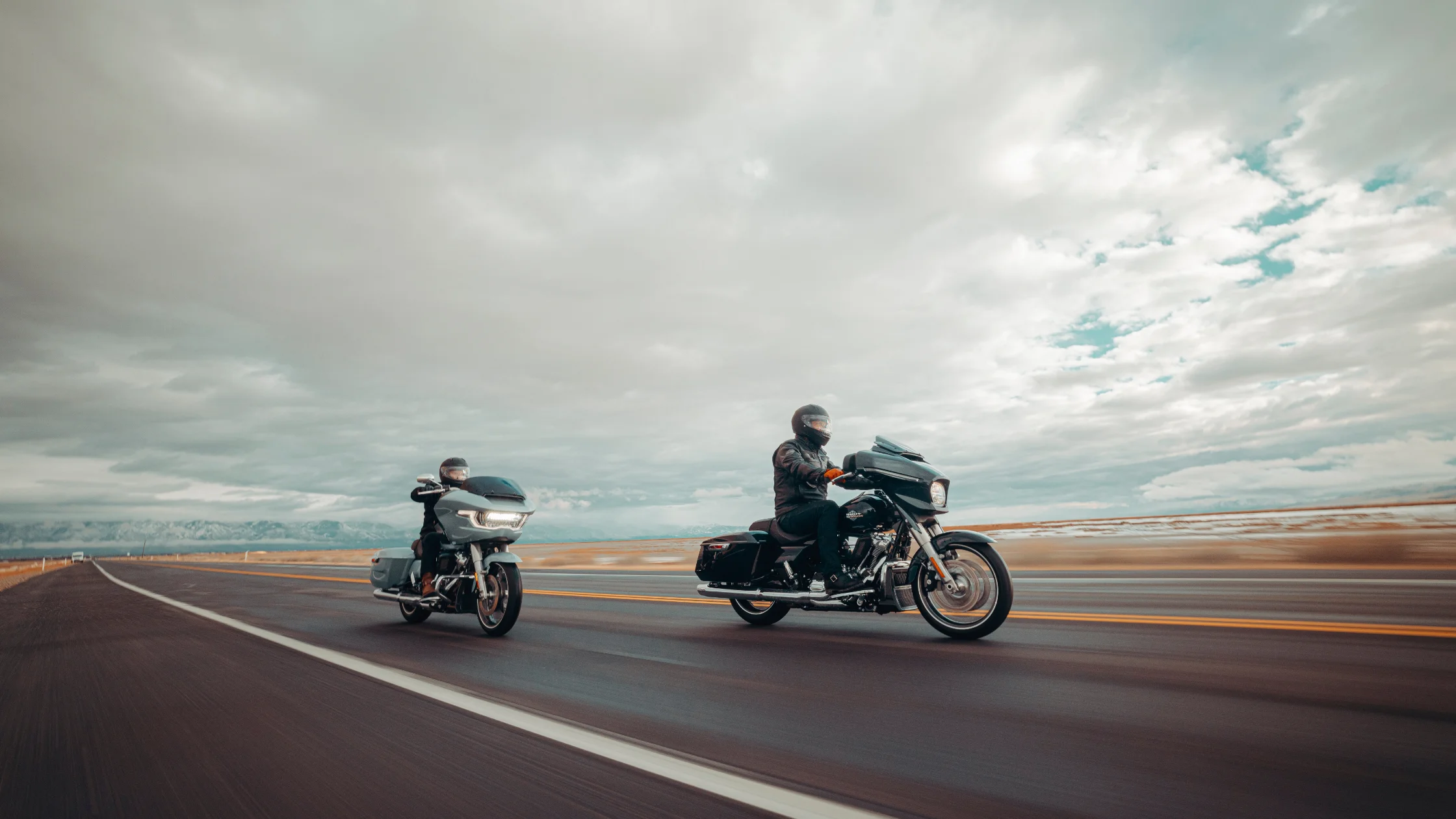 2025 Harley-Davidson Street Glide and Road Glide cruising side by side on an open highway under a dramatic cloudy sky, showcasing their touring capabilities and sleek aerodynamic design.