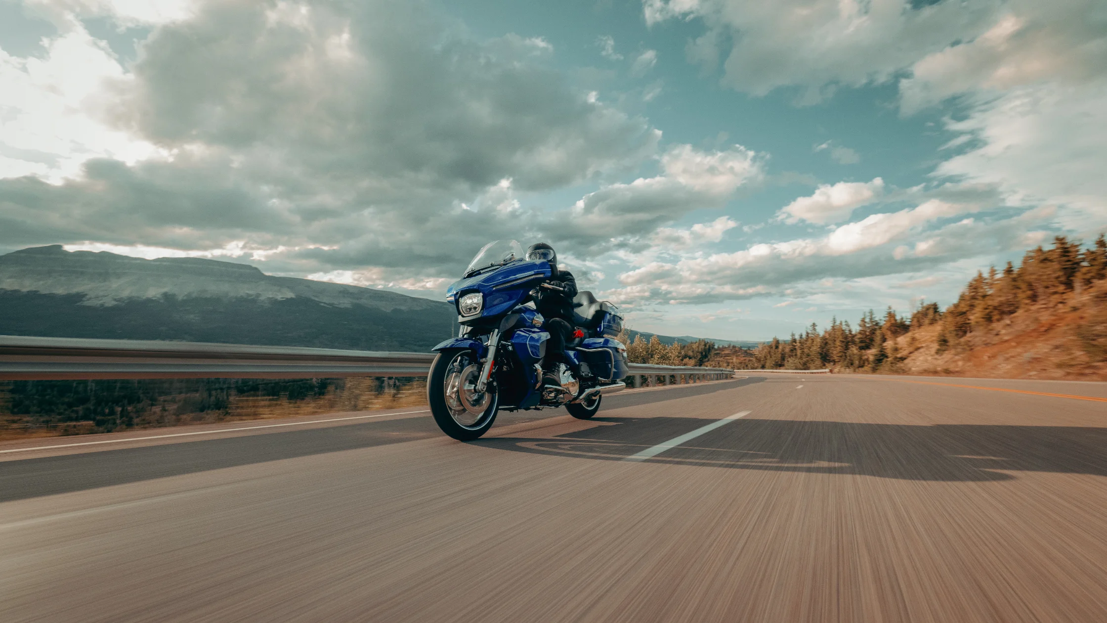 2025 Harley-Davidson Street Glide Ultra in deep blue, cruising along an open highway with a scenic mountain backdrop, showcasing its premium touring features and comfortable long-distance capability.