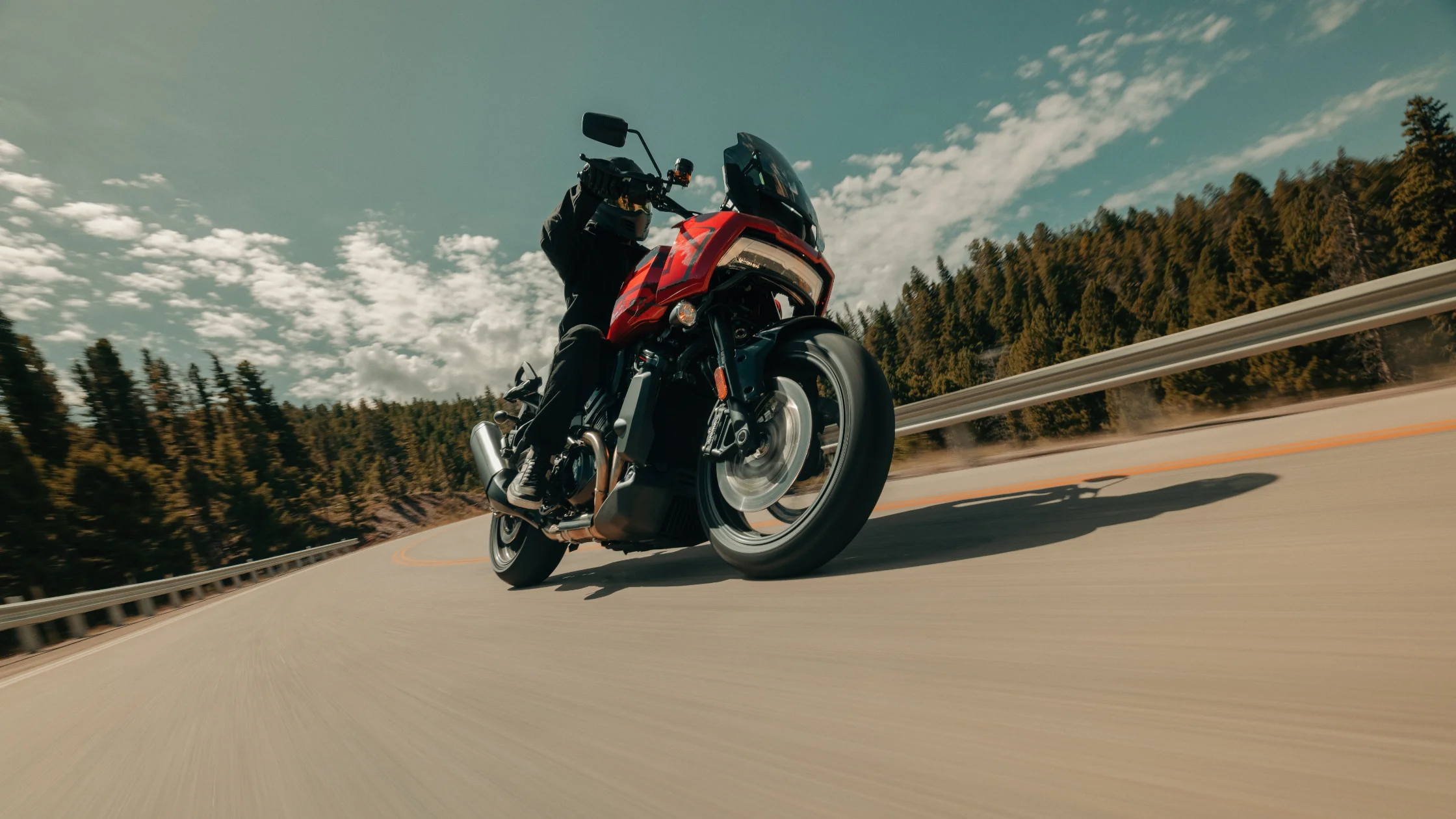 2025 Harley-Davidson Pan America 1250 Special in vibrant red, leaning aggressively into a curve on a scenic highway, highlighting its adventure touring prowess and rugged design.