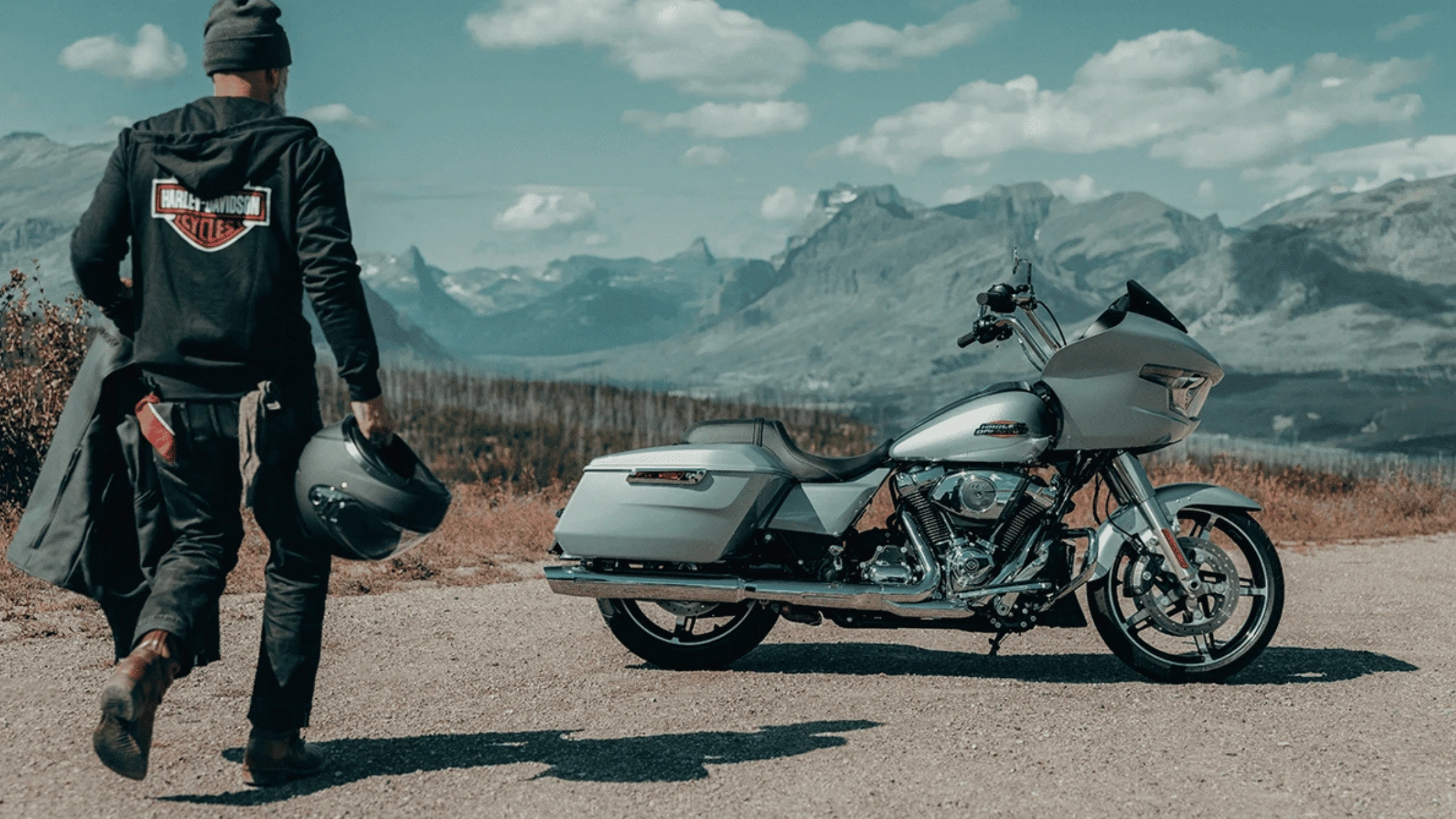 Biker walking toward a silver Harley-Davidson Road Glide motorcycle parked with a scenic mountain backdrop.