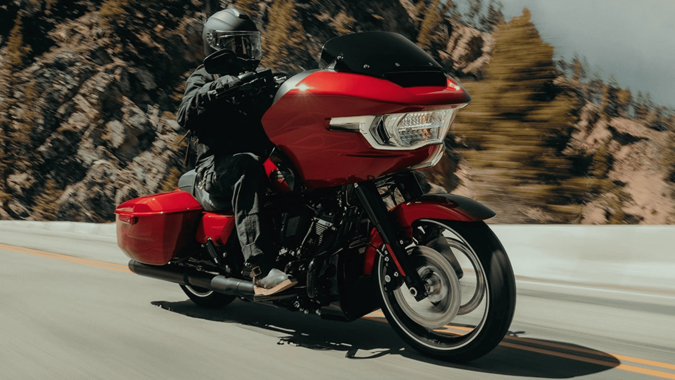 A rider on a red 2025 Harley-Davidson® Road Glide® leans into a curve on a scenic mountain highway.