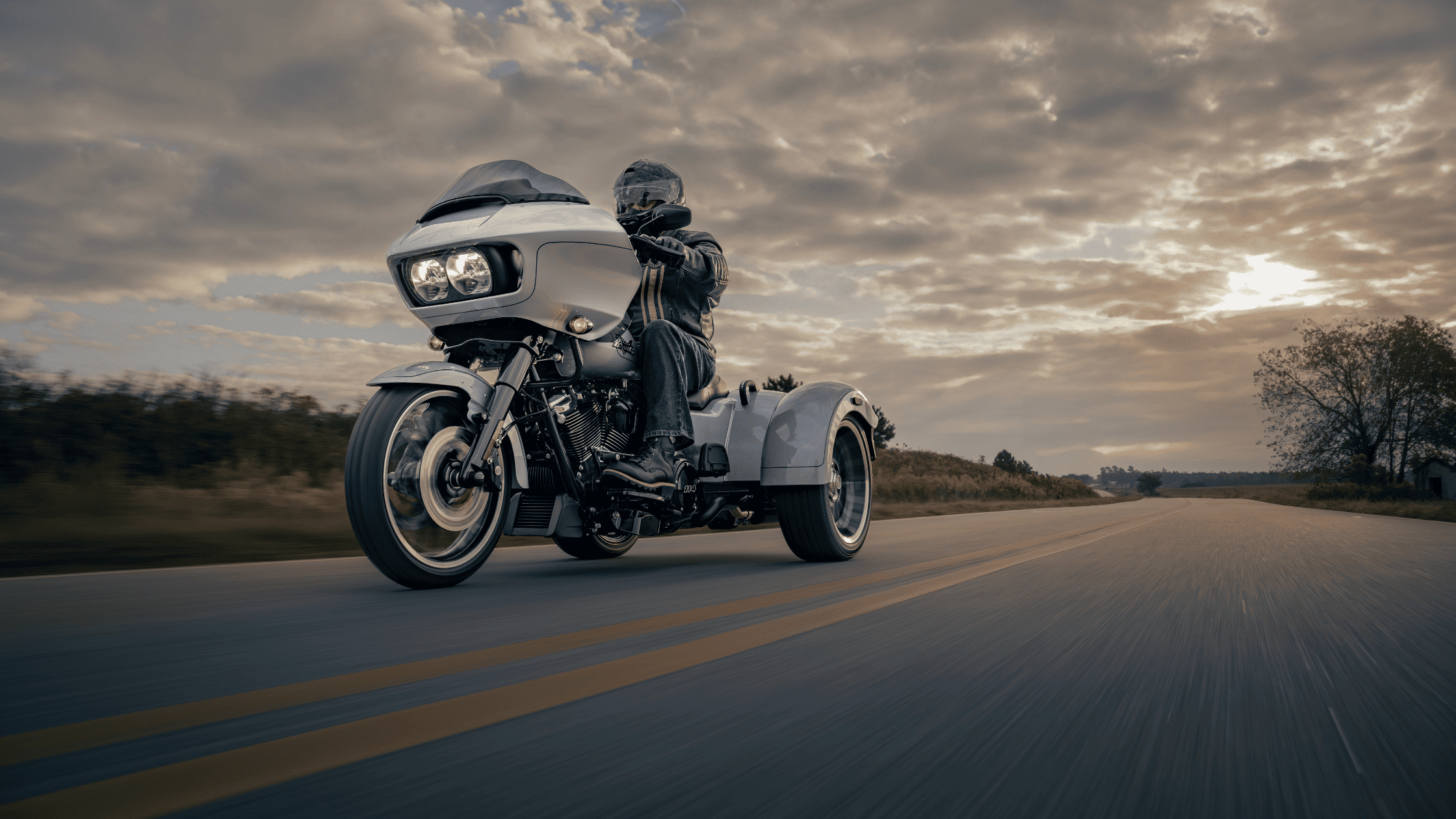 A rider cruises on a 2025 Harley-Davidson® Road Glide® 3 trike down an open highway under a dramatic sky.