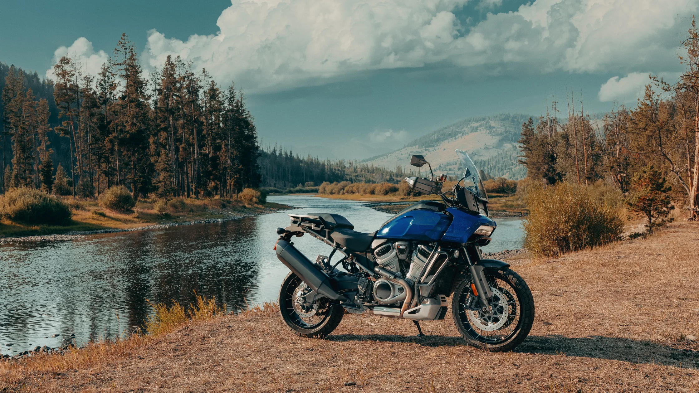 2025 Pan America 1250 S parked riverside with pine trees and mountains in the background.