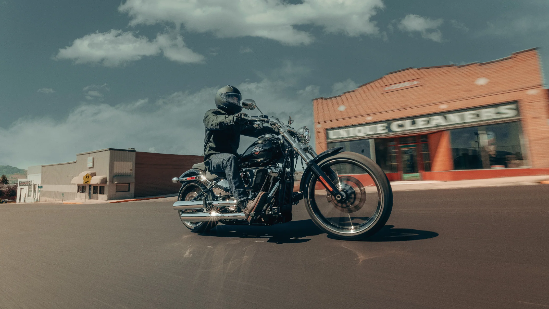 2025 Harley-Davidson® Breakout® rider cruising past brick Unique Cleaners building on sunny day.