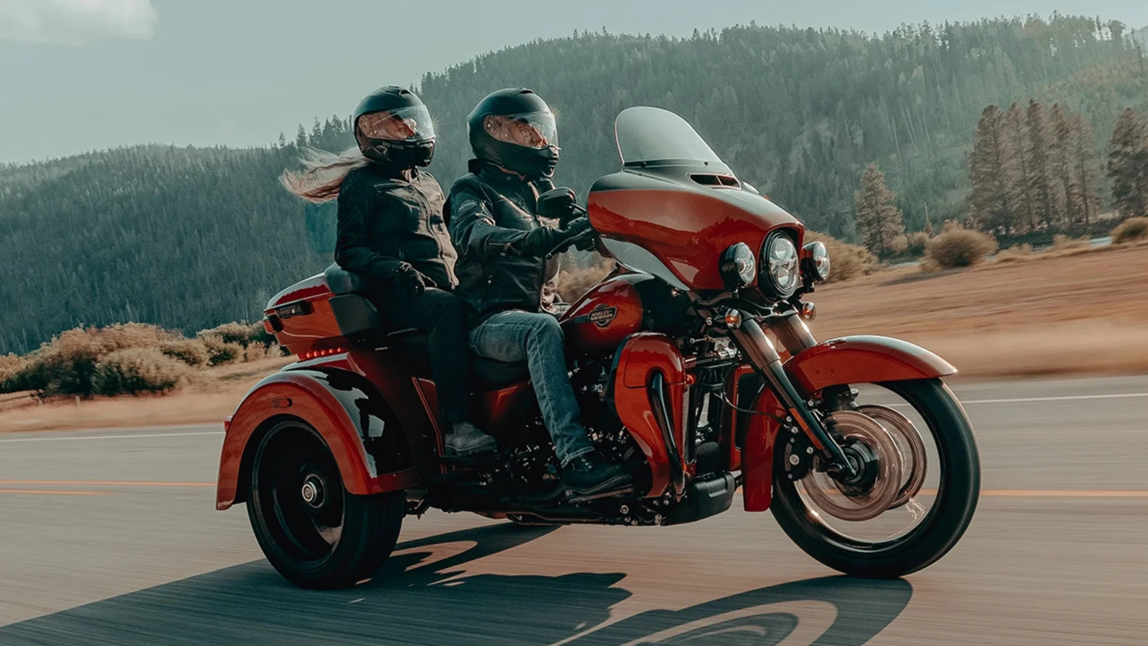 Two riders on a 2025 Harley-Davidson® Tri Glide® Ultra cruising past forested hills on open road.