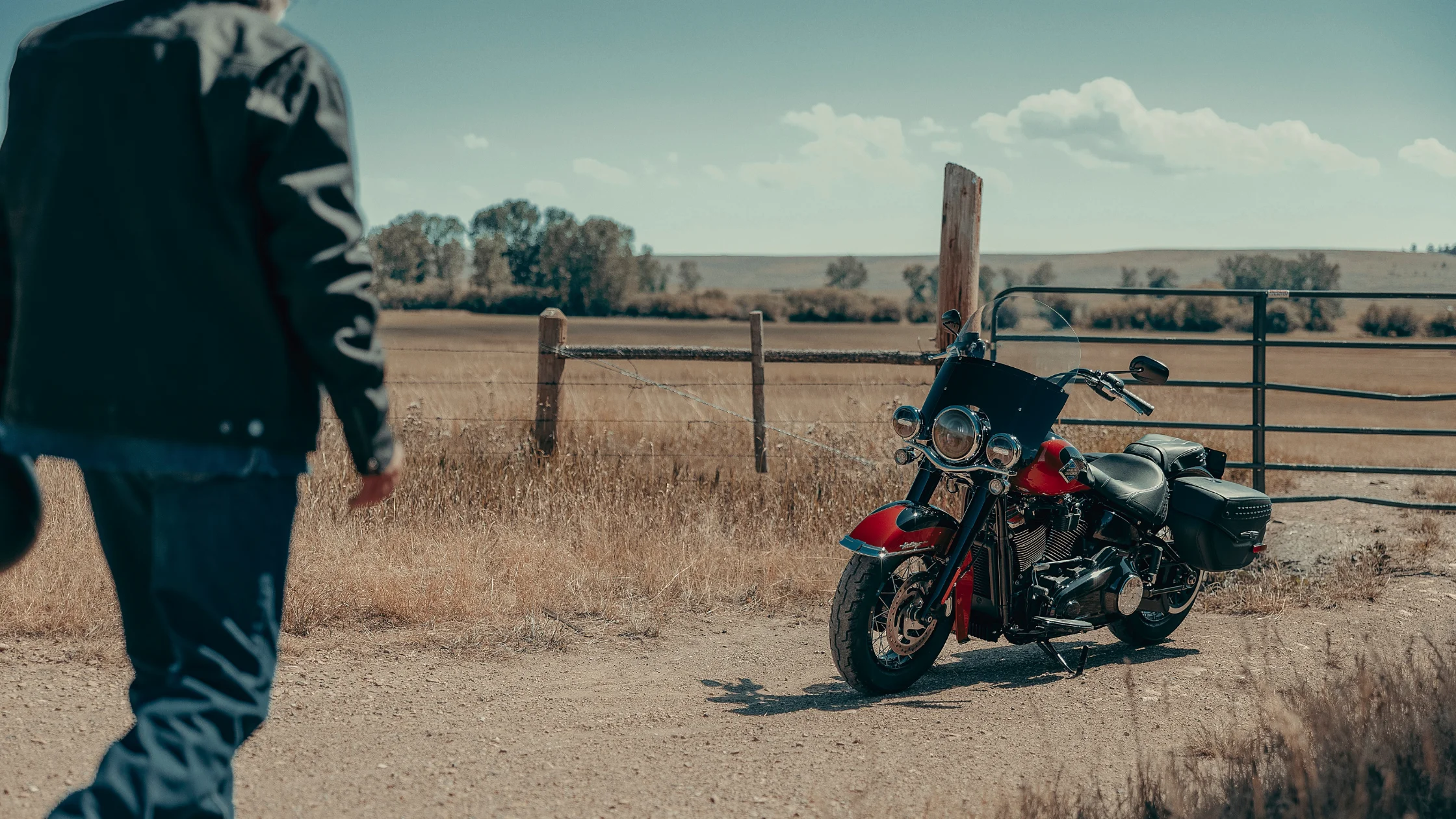 2025 Harley-Davidson® Heritage Classic parked near a fence in a rural field as rider approaches.