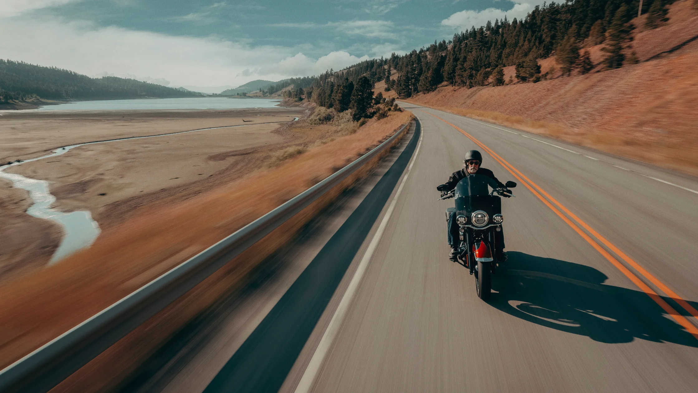 2025 Harley-Davidson® Heritage Classic riding along a scenic highway with lake and mountain backdrop.