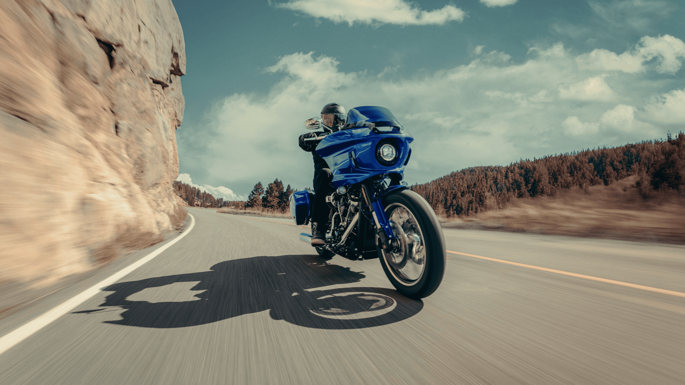 2025 Harley-Davidson Low Rider ST in bright blue riding past rock cliffs and pine forest.