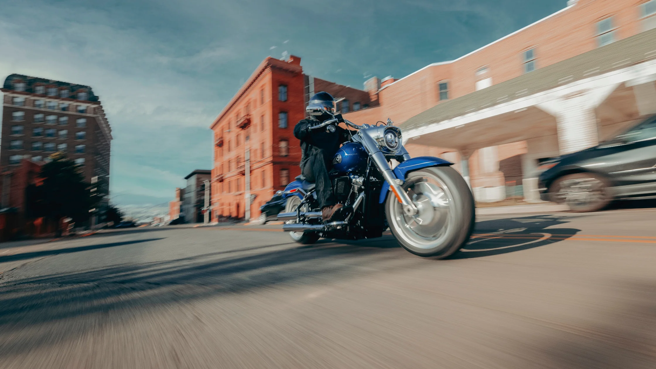 2025 Harley-Davidson Fat Boy in vivid blue, cruising through an urban street with vintage brick buildings.