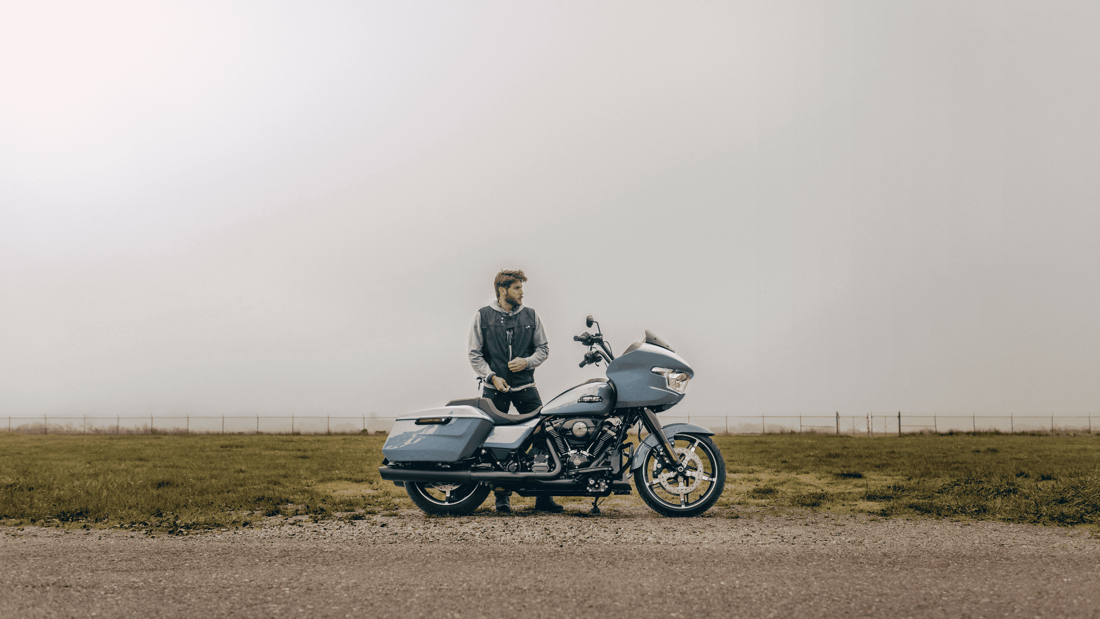 Man standing next to a blue 2025 Harley-Davidson Road Glide on a misty open road with grassy fields