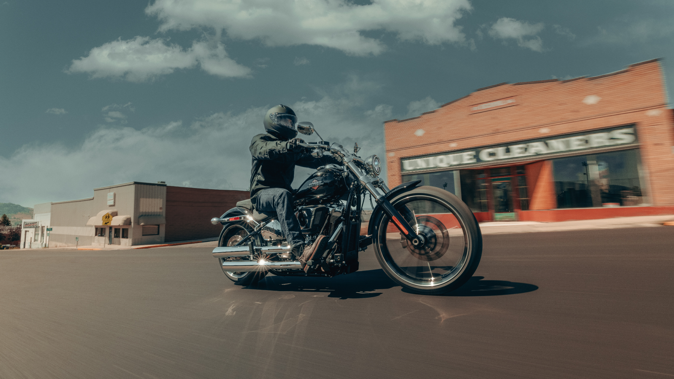 Rider on a 2025 Harley-Davidson® Breakout® cruising through a small-town street, passing brick and storefront buildings under a bright blue sky.