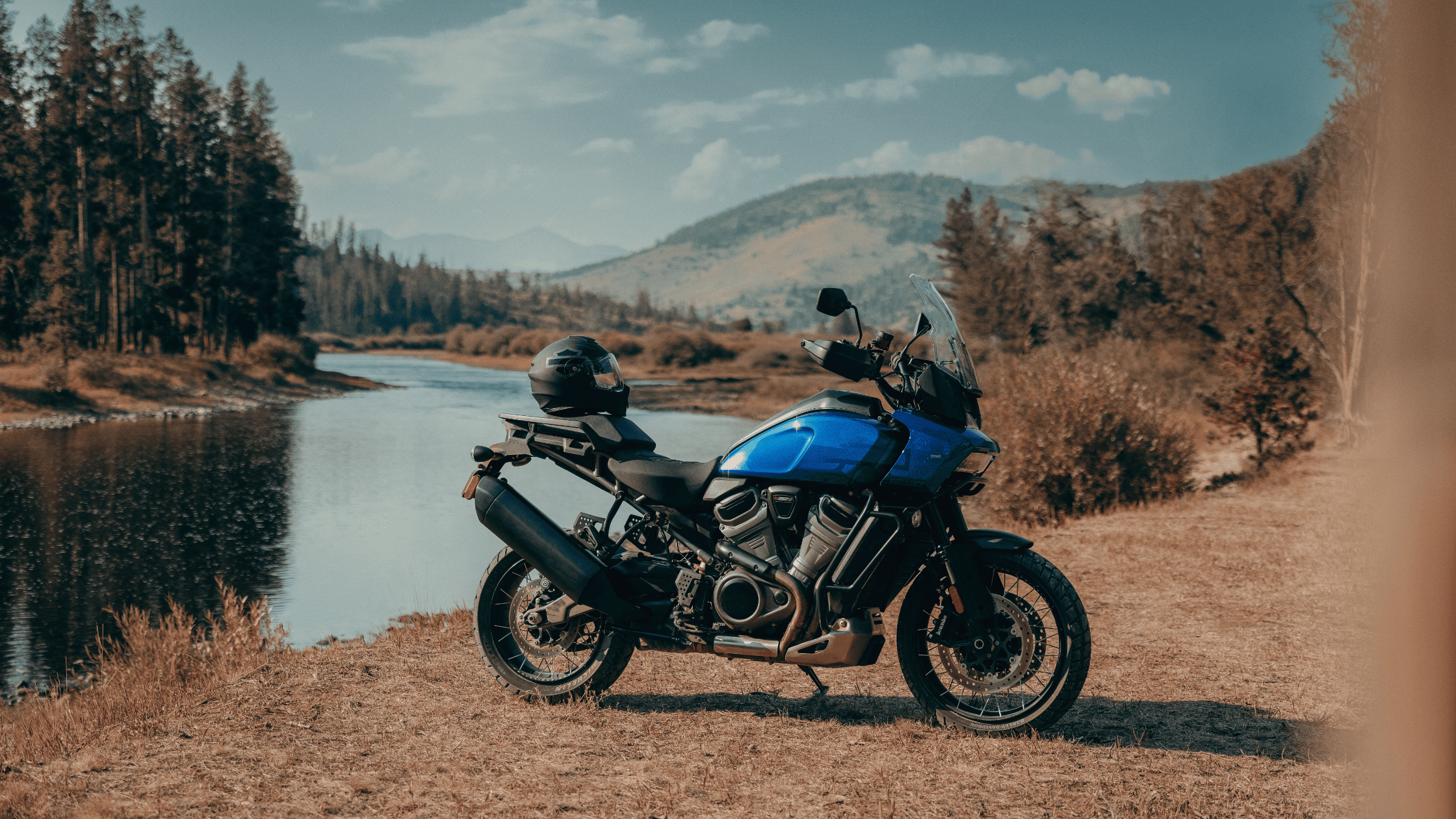 Blue 2025 Harley-Davidson Pan America 1250 Special parked riverside with mountains and pine forest in the background.