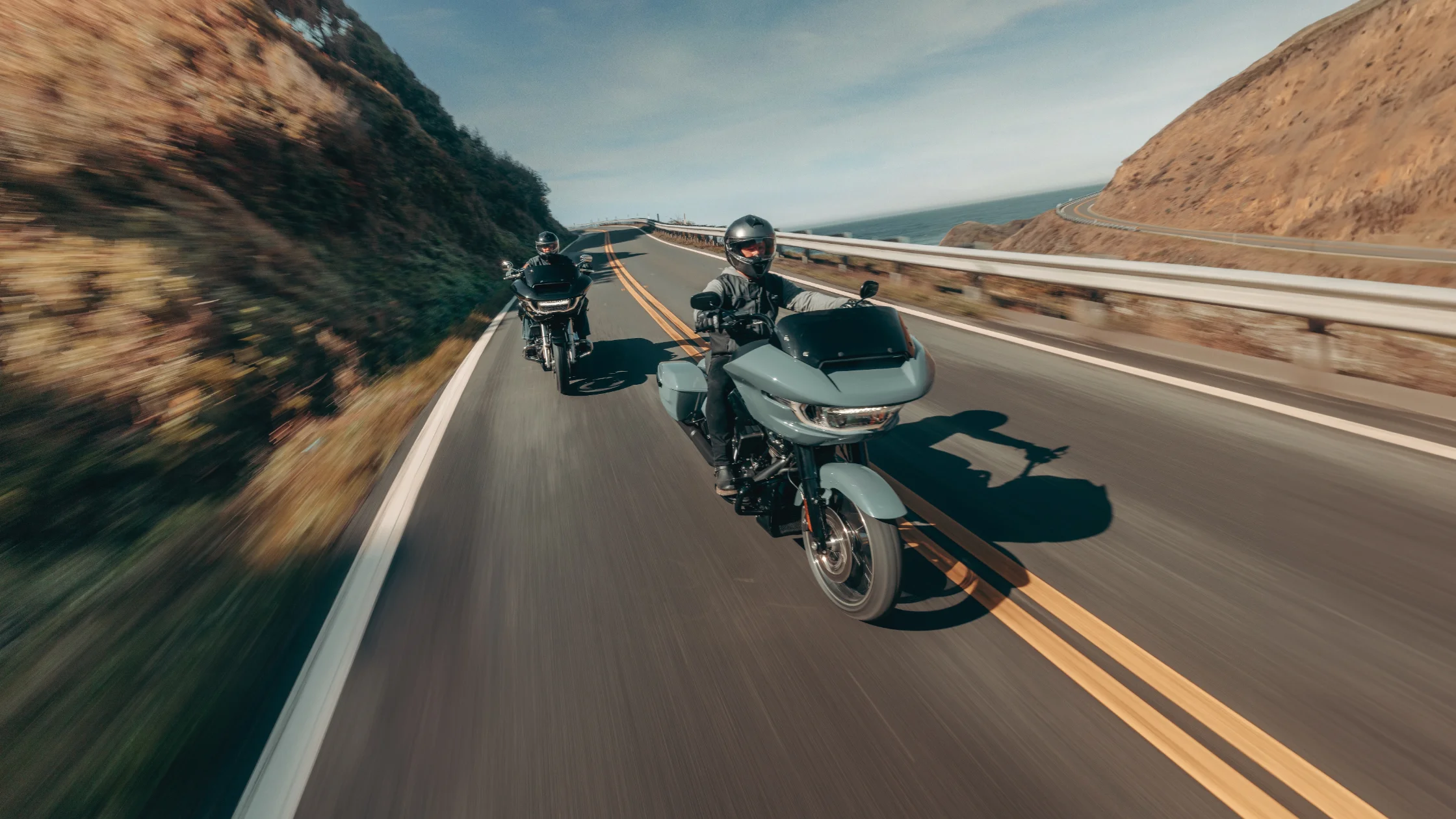 2025 H-D® Road Glide® Carryover in matte blue leading a second bike on a coastal highway, overhead trailing angle