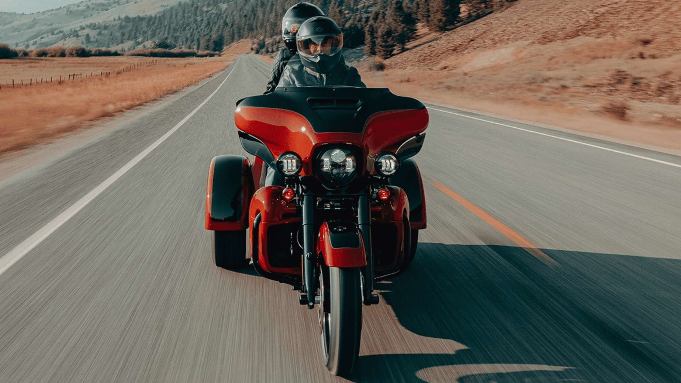 Two-up riders on a 2025 Harley-Davidson® Tri Glide® Ultra in striking red finish gliding down a scenic highway near Atlanta, GA.
