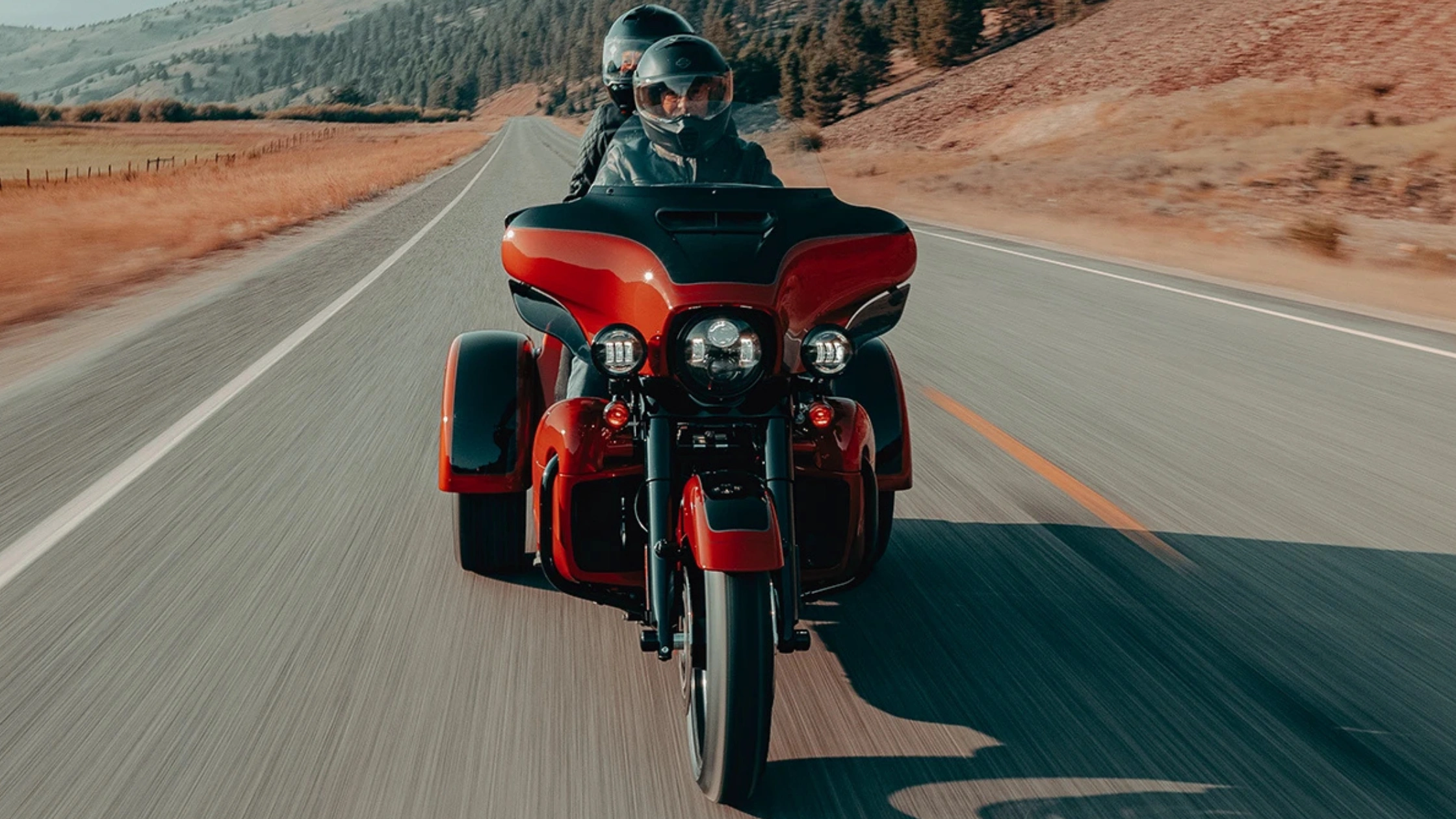 Two-up riding on 2025 Tri Glide Ultra Carryover in red and black, moving along a rural highway.