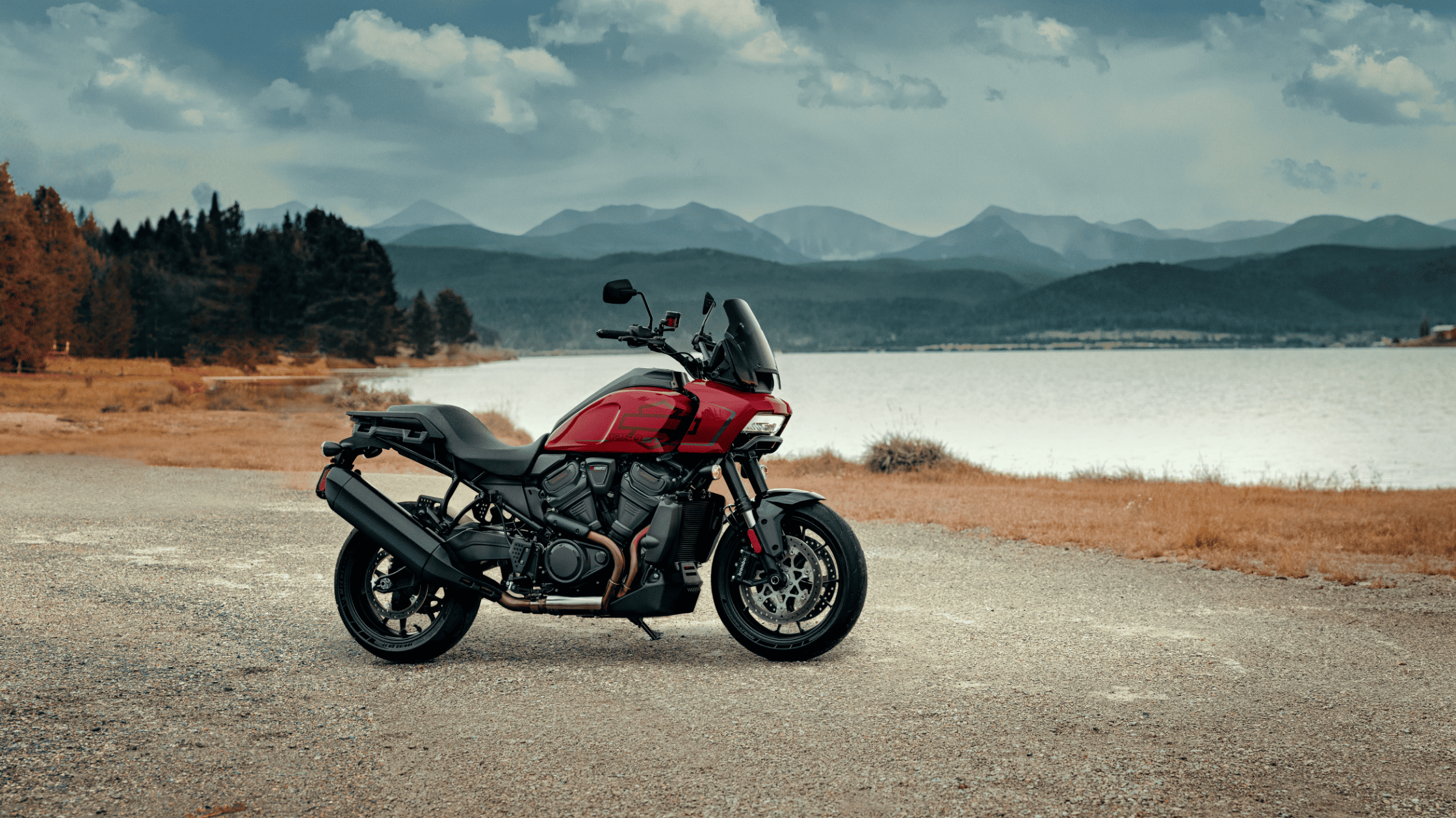2025 Pan America 1250 ST in red parked lakeside with mountain range under cloudy sky