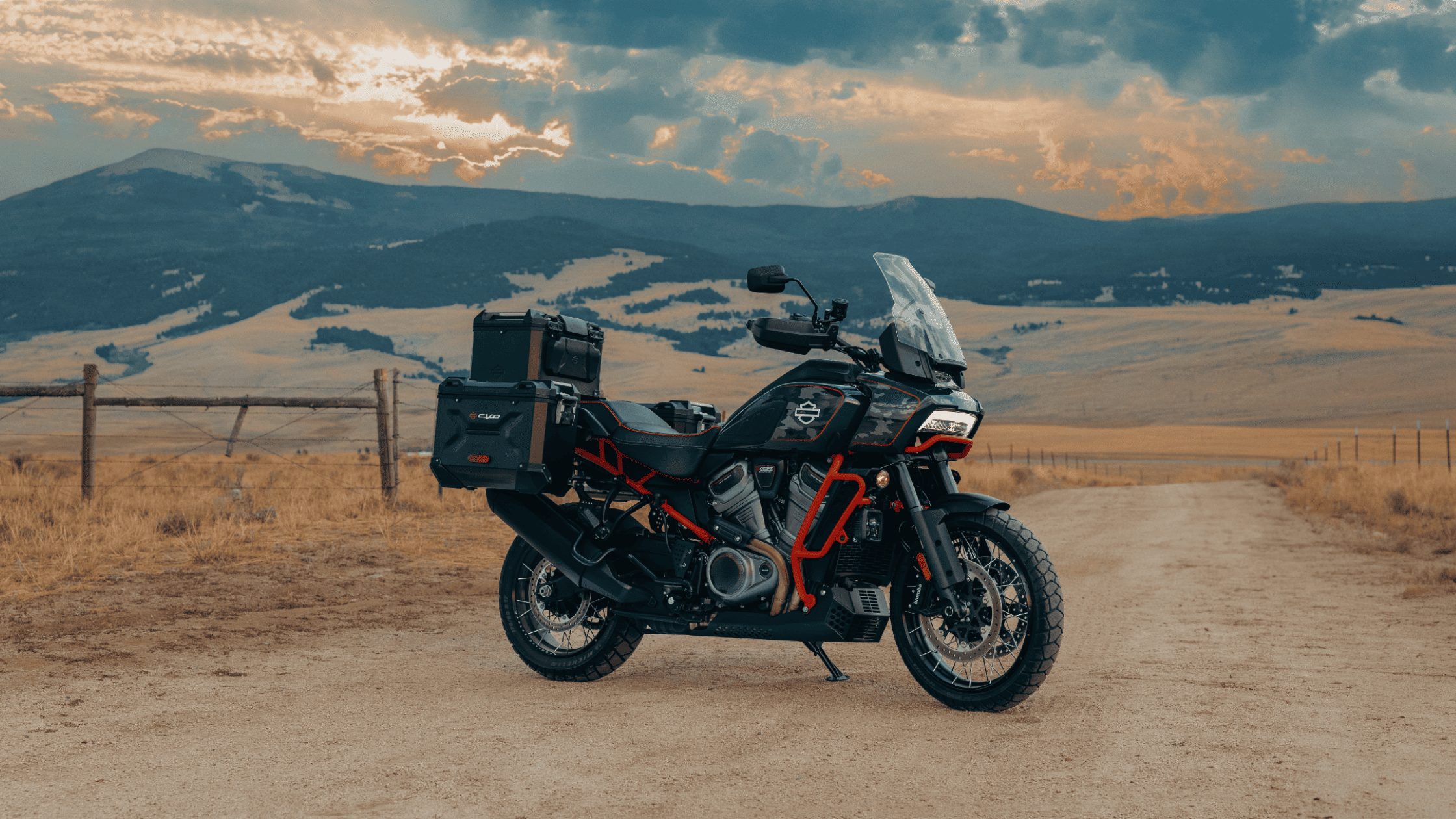 A 2025 Harley-Davidson® CVO™ Pan America® adventure touring motorcycle stands on a dirt trail with mountains in the distance, highlighting its rugged off-road capability, tall windshield, and side panniers under a vibrant sunset sky.