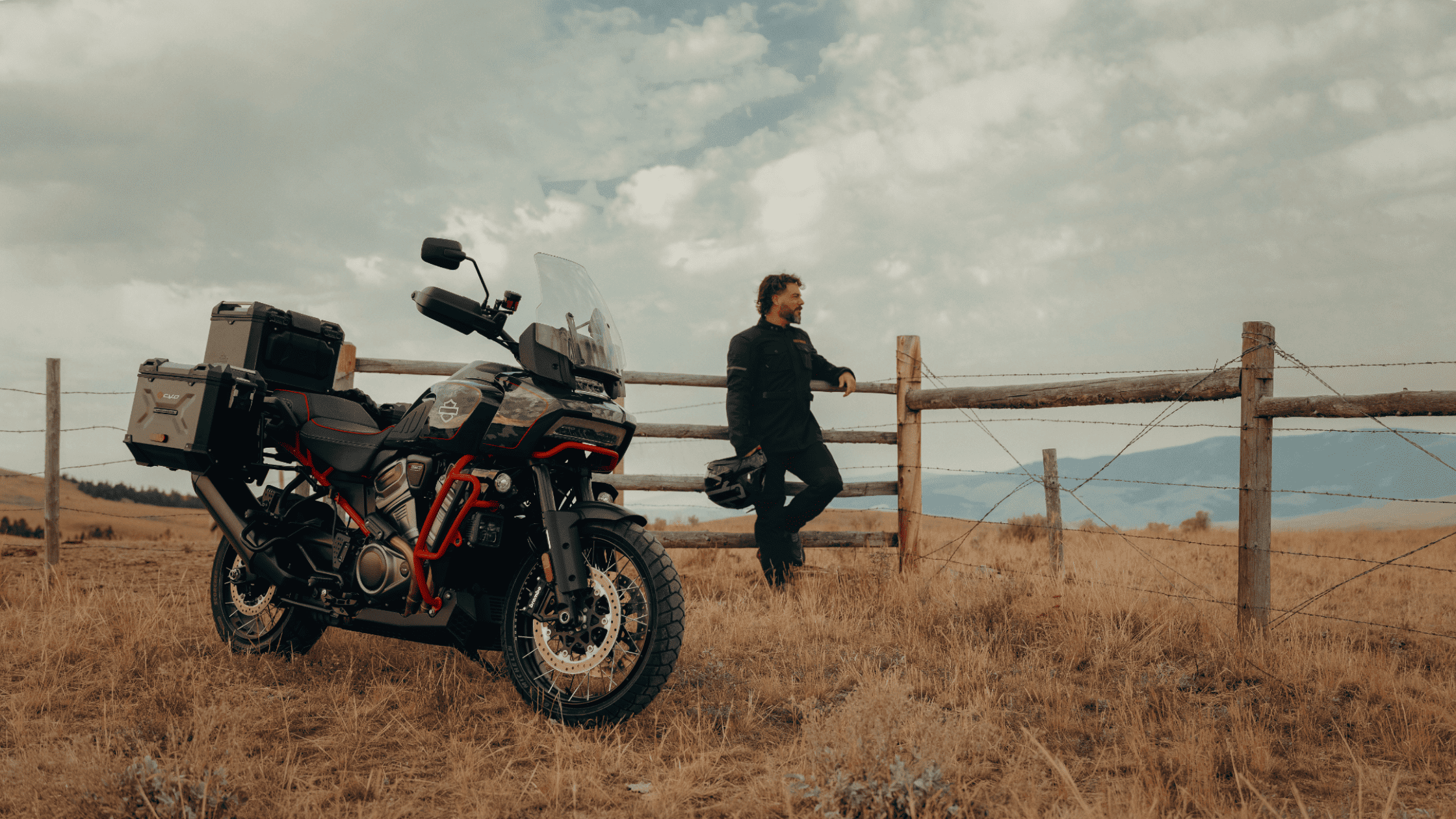 A 2025 Harley-Davidson® CVO™ Pan America® adventure motorcycle parked beside a rustic wooden fence, with a rider standing nearby under a cloudy sky. The rugged landscape emphasizes its off-road touring capability and adventure-ready design.