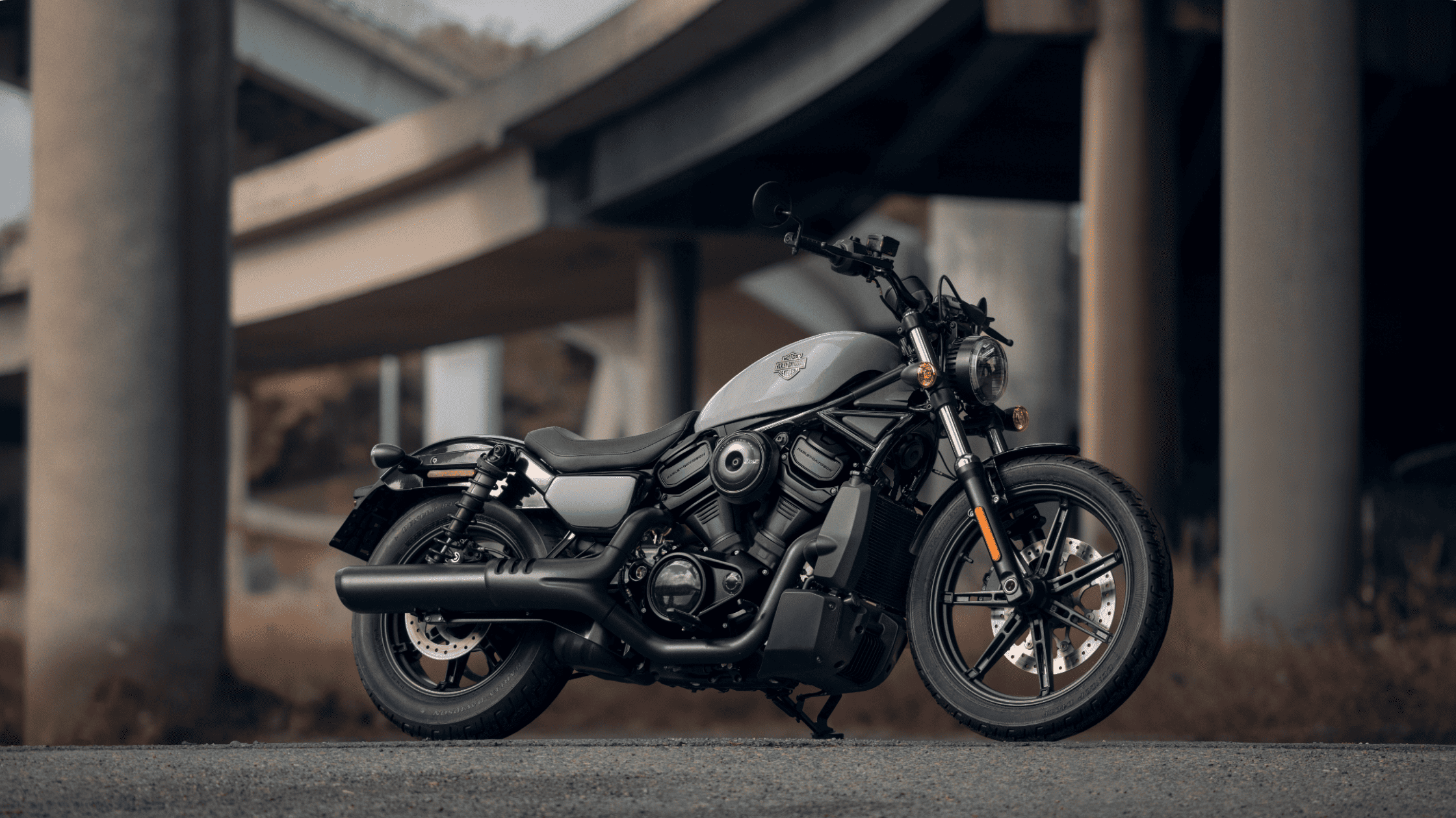 A 2025 Harley-Davidson® Nightster® cruiser rests under an overpass, featuring a sleek silver tank, low profile, and blacked-out exhaust system designed for agile urban riding.