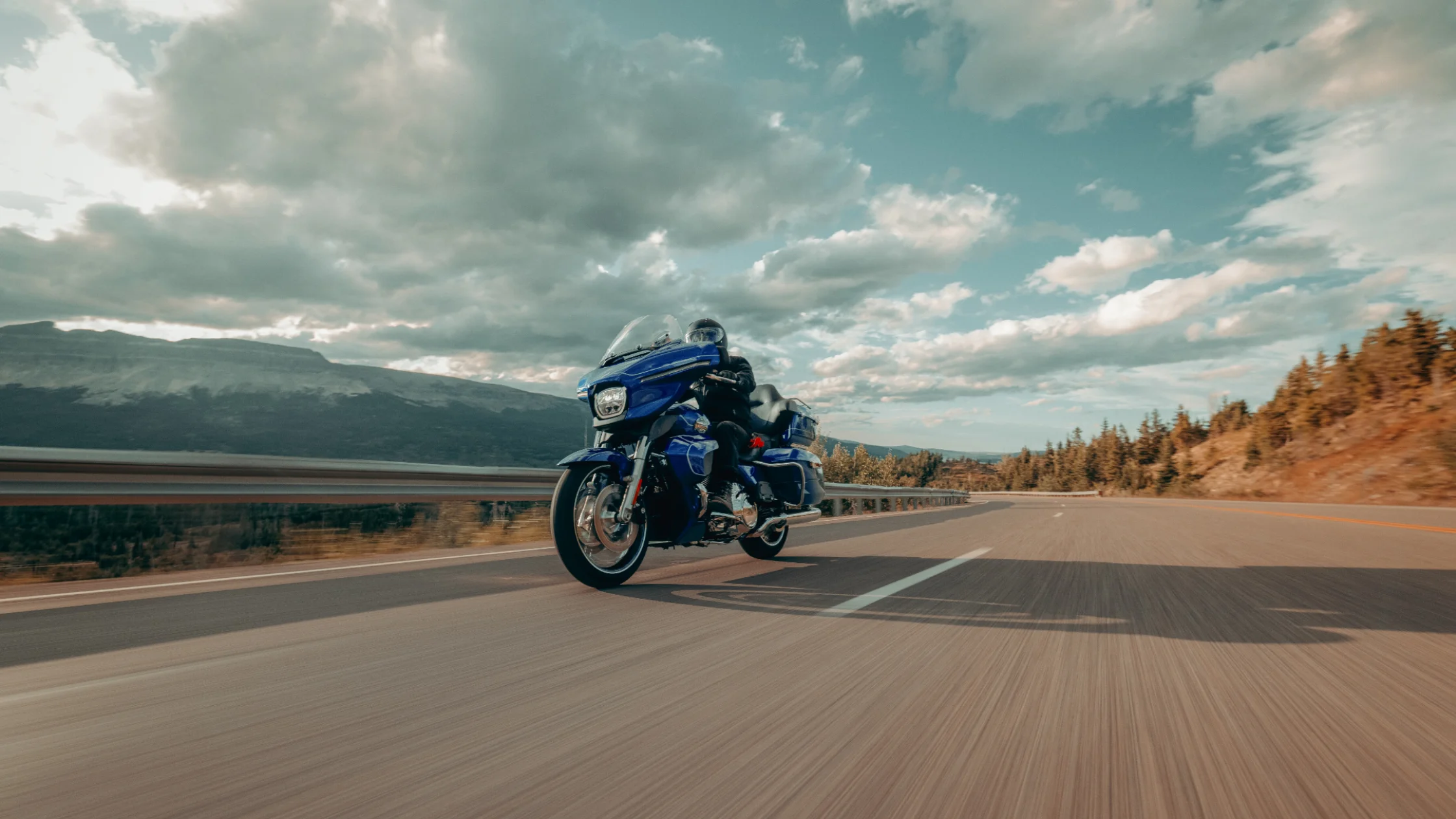 Blue 2025 Harley-Davidson Street Glide Ultra cruising a mountain highway beneath bright afternoon clouds.