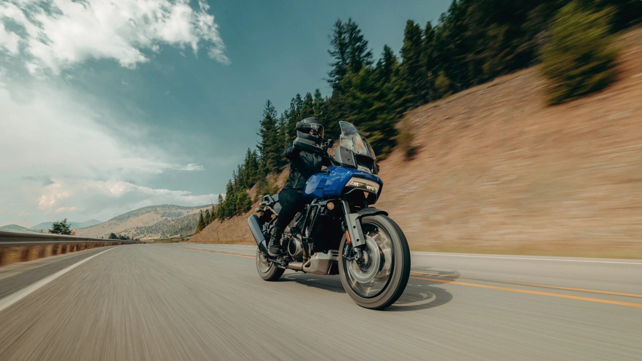 Rider on a blue 2025 Harley-Davidson Pan America 1250 Special on a highway surrounded by forested hills.
