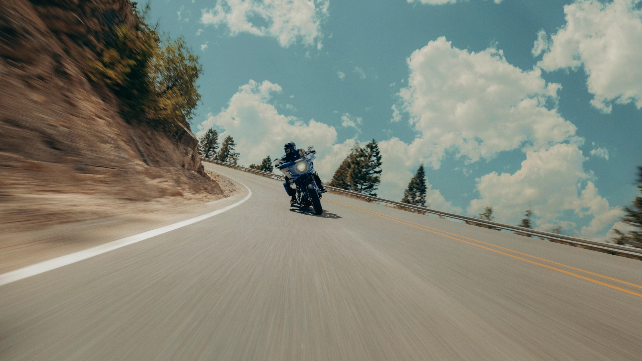 2025 Harley-Davidson® Low Rider® ST leaning into a scenic mountain curve on a sunny day with clouds overhead.