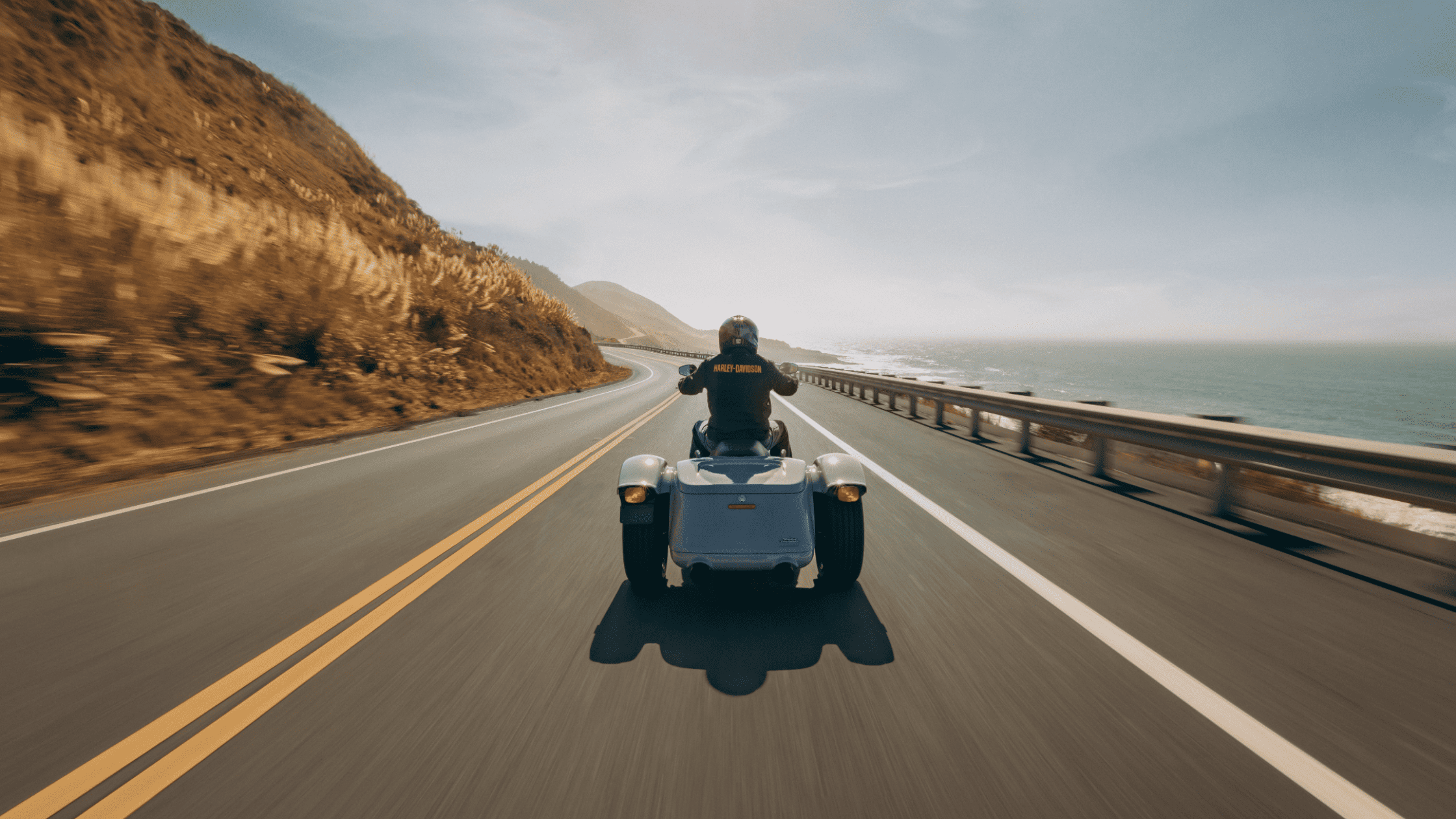 2025 Harley-Davidson Freewheeler trike cruising a coastal highway with rider in full gear from rear view