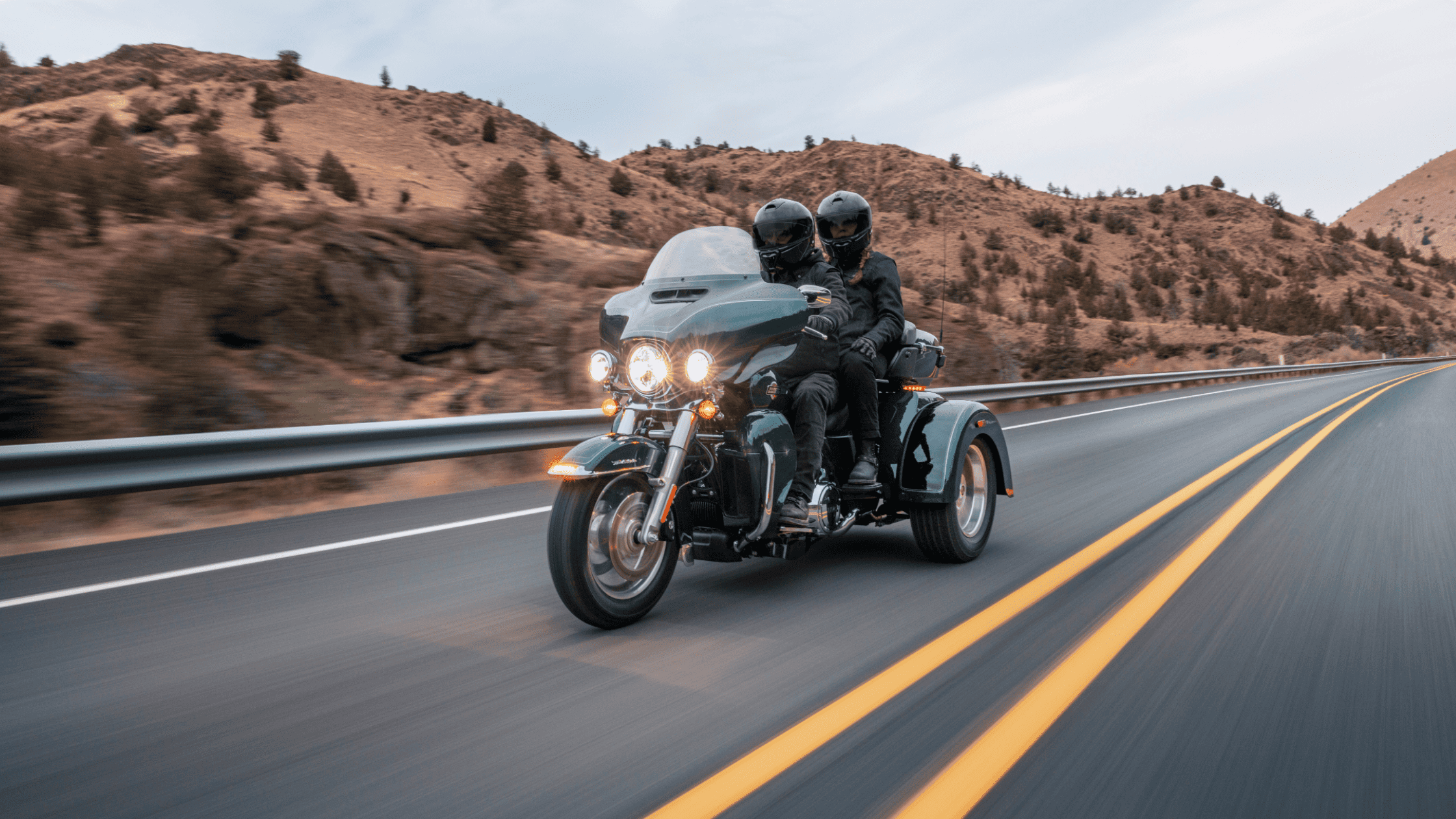 Two riders on a 2025 Harley-Davidson® Tri Glide® Ultra touring trike on an open highway, capturing its long-distance comfort and luxury touring features.