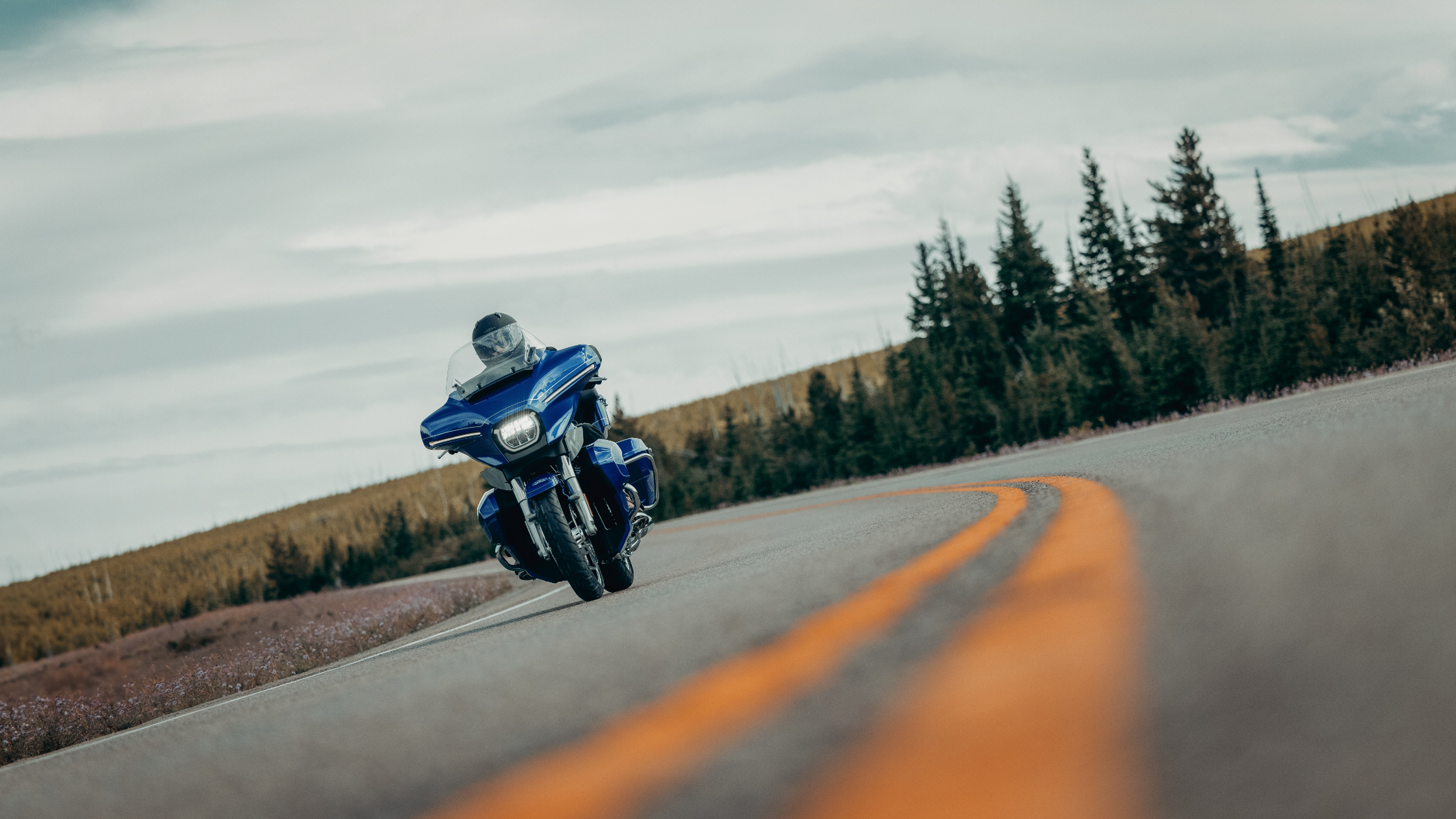 2025 Harley-Davidson® Street Glide® Ultra in vivid blue cruising along a winding mountain highway surrounded by trees.