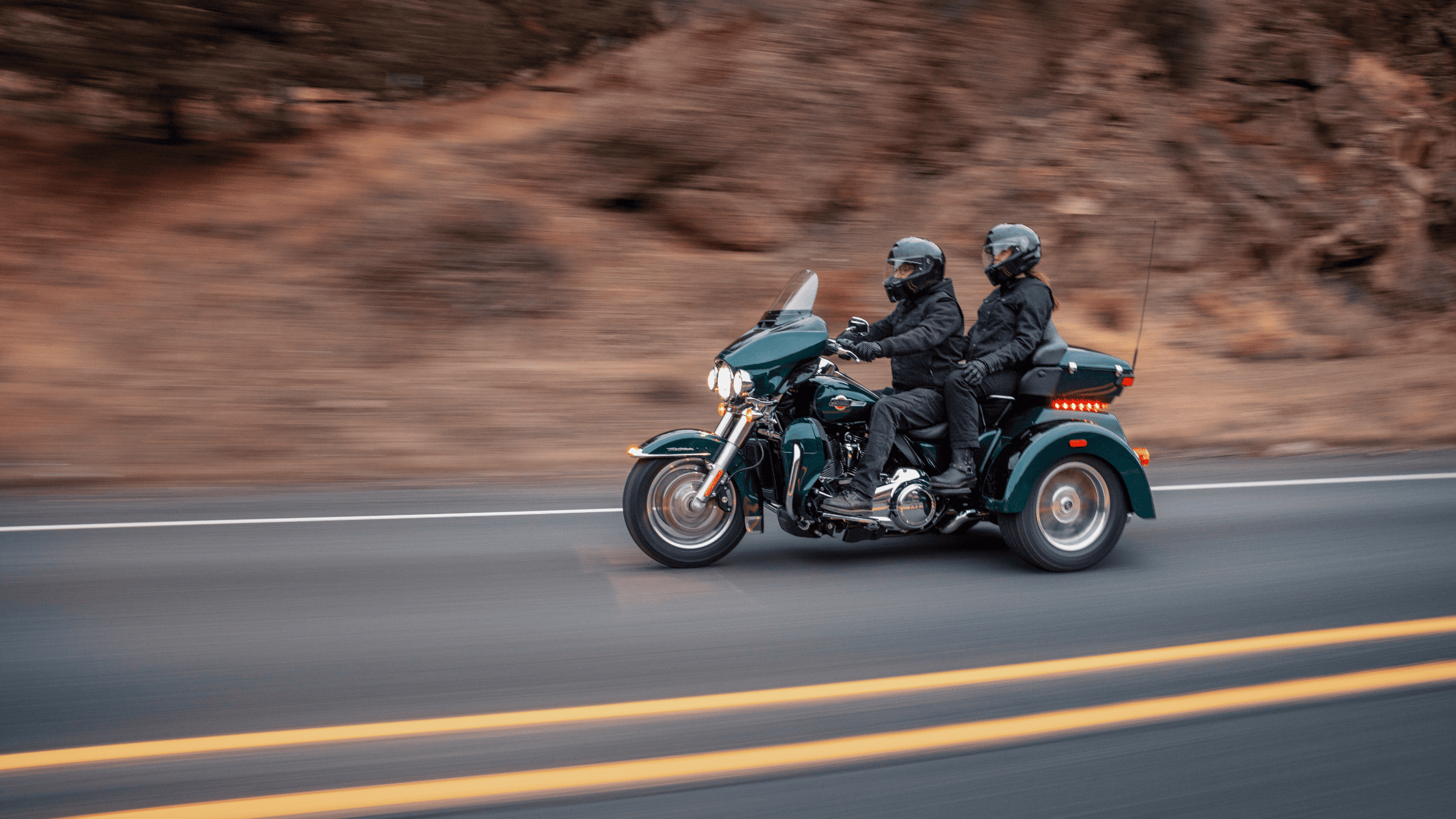 Couple riding a 2025 Harley-Davidson® Tri Glide® Ultra in dark green on a winding road with blurred rocky backdrop.